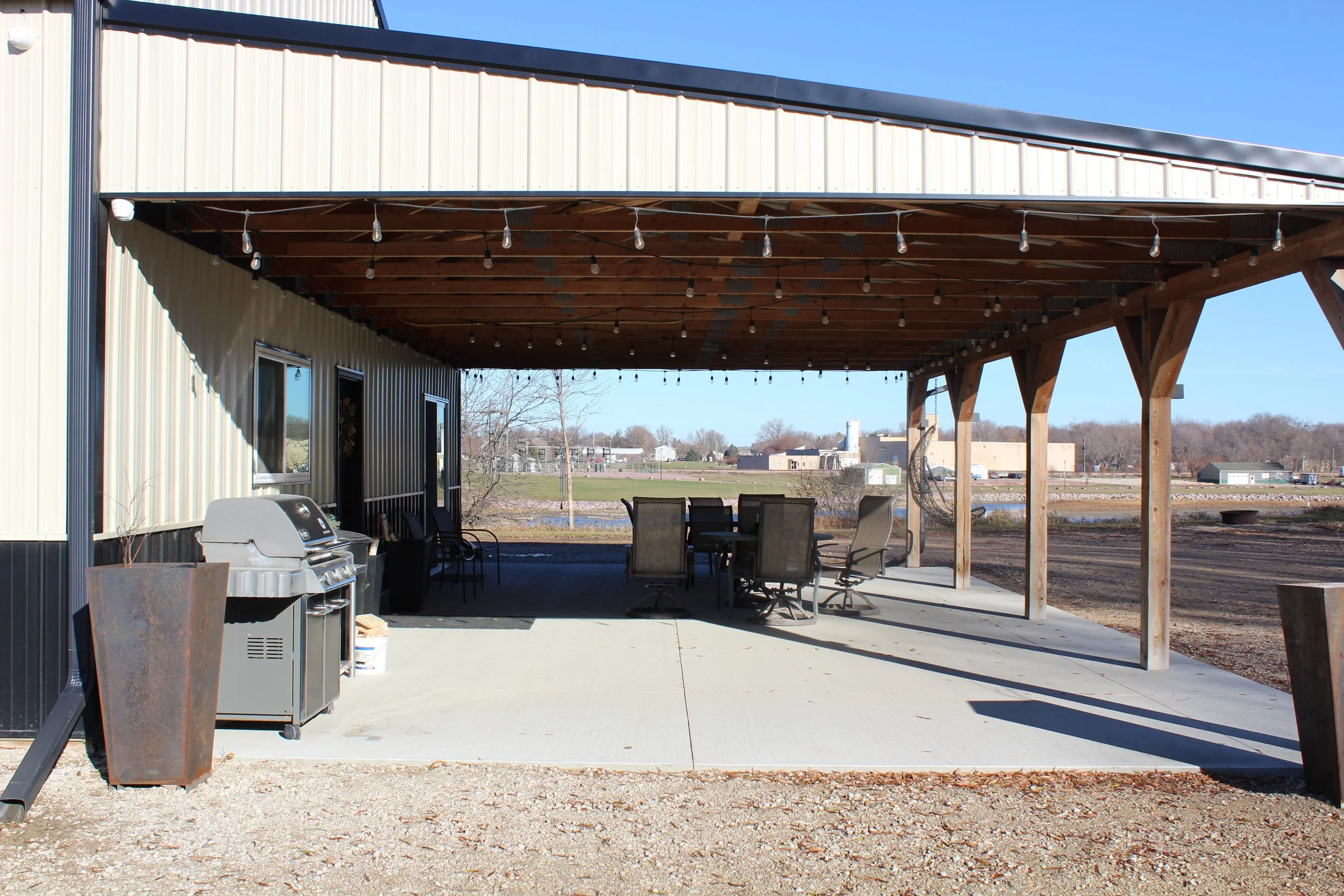 Outdoor patio area with a grilling station, chairs, a table, and hanging lights, attached to a building with a metal exterior, overlooking a rural landscape with a pond and trees.