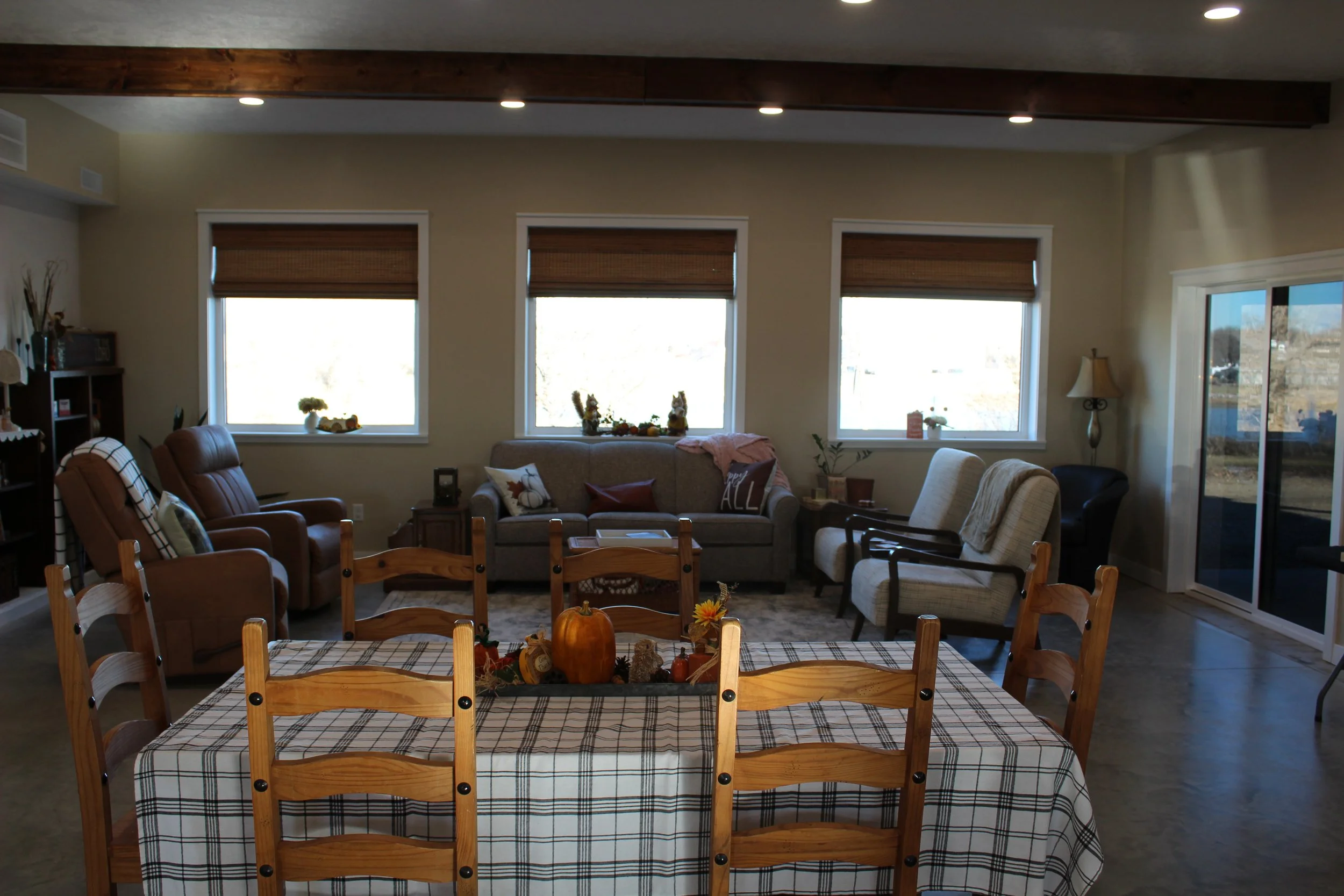 Living room with wooden beams, large windows, and a sliding glass door, furnished with chairs, sofas, a dining table with a checkered tablecloth, and autumn-themed decorations including pumpkins.