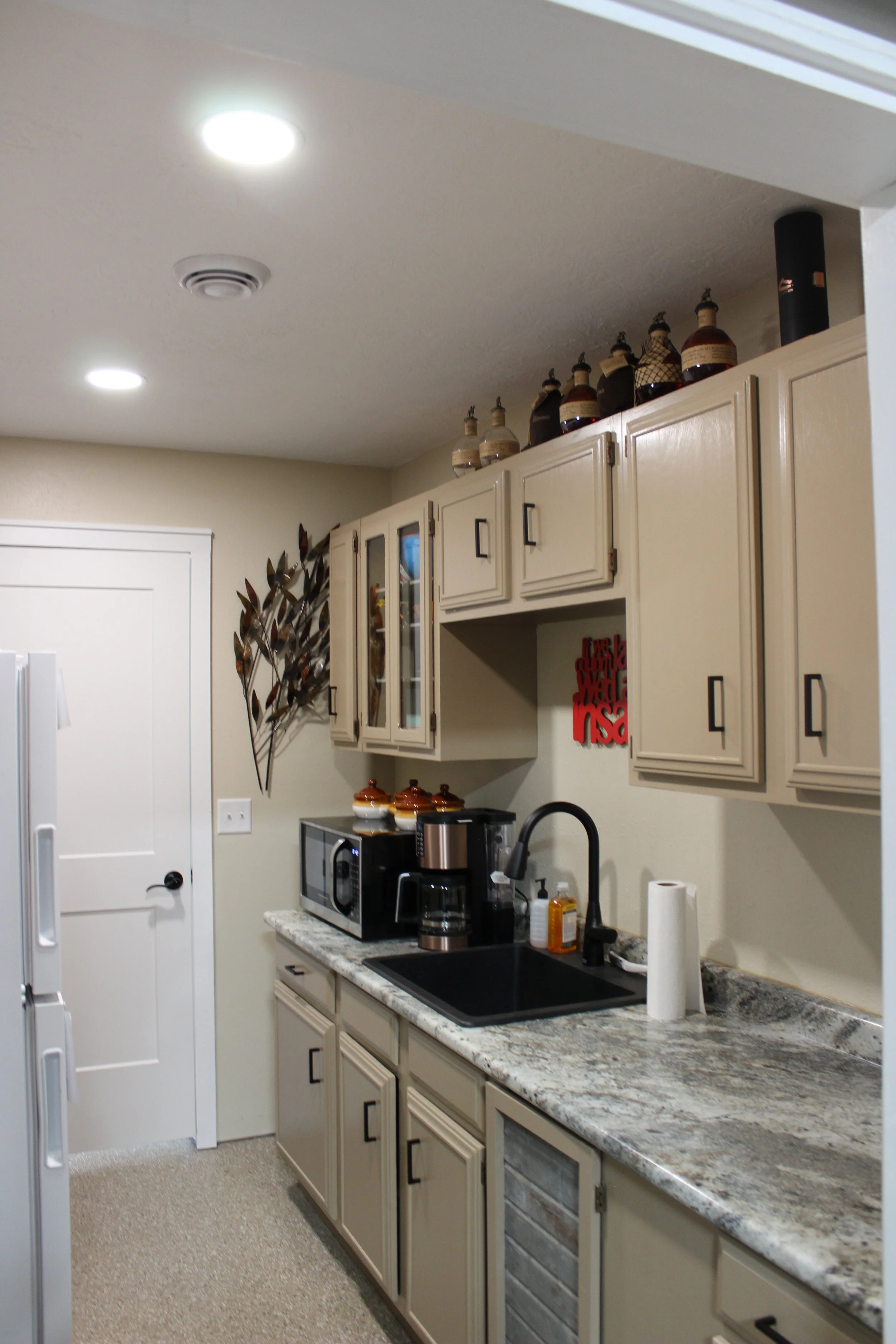 A kitchen with beige cabinets, granite countertops, a black sink, and various appliances including a coffee maker, microwave, and toasters. Decorative bottles are on top of the cabinets, and there is a wall decoration on the right side.