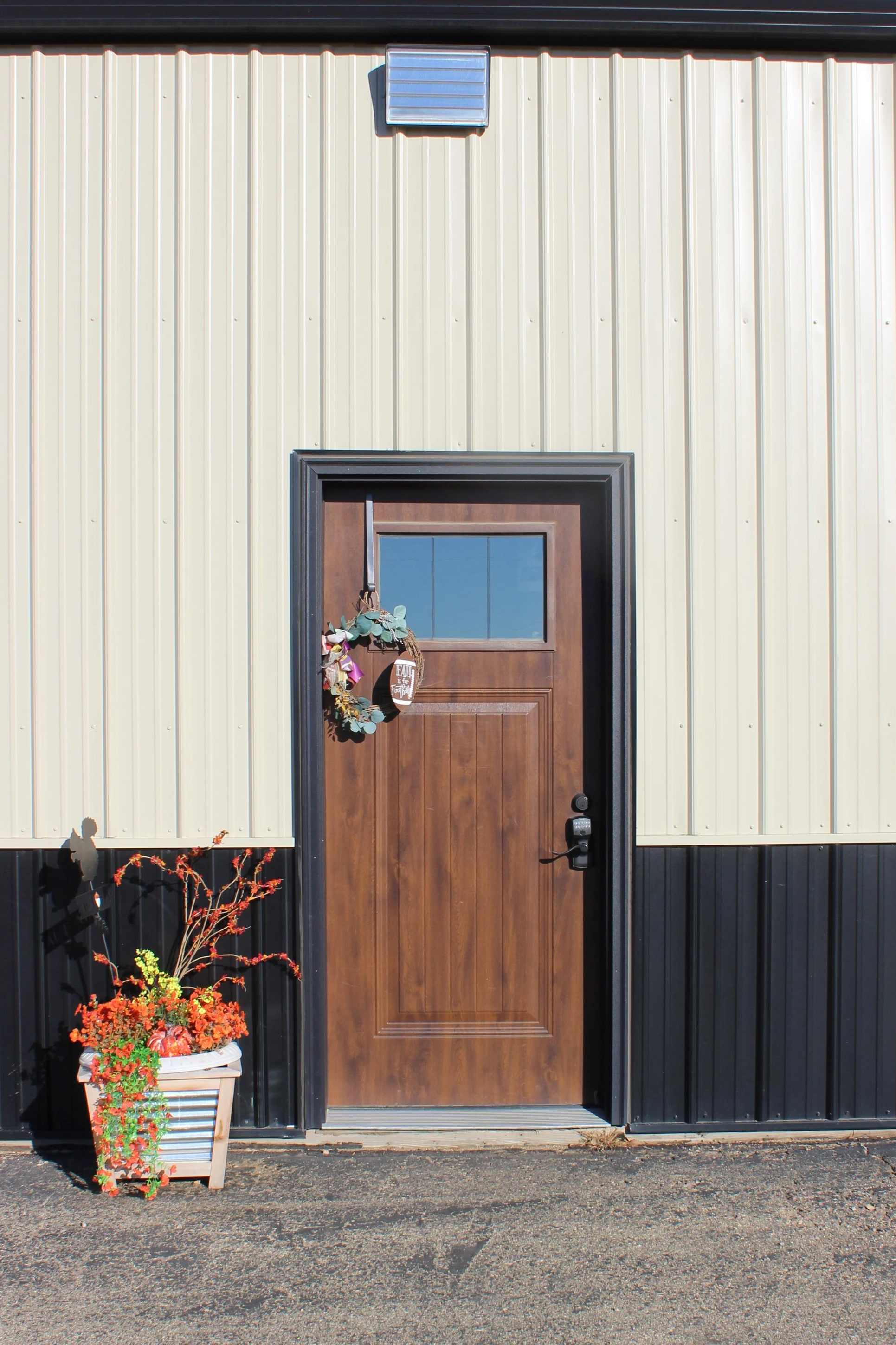 Exterior view of a building entrance with a wooden door, a decorative wreath hanging on the door, a window at the top of the door, a flower pot with orange flowers, and black and beige corrugated metal walls.