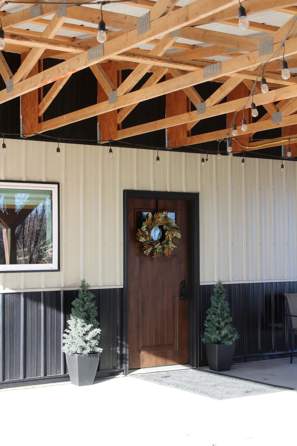 Wooden porch ceiling with exposed beams and string lights. Brown door with Christmas wreath, flanked by two potted pine trees. Part of a window on the left and outdoor furniture on the right.
