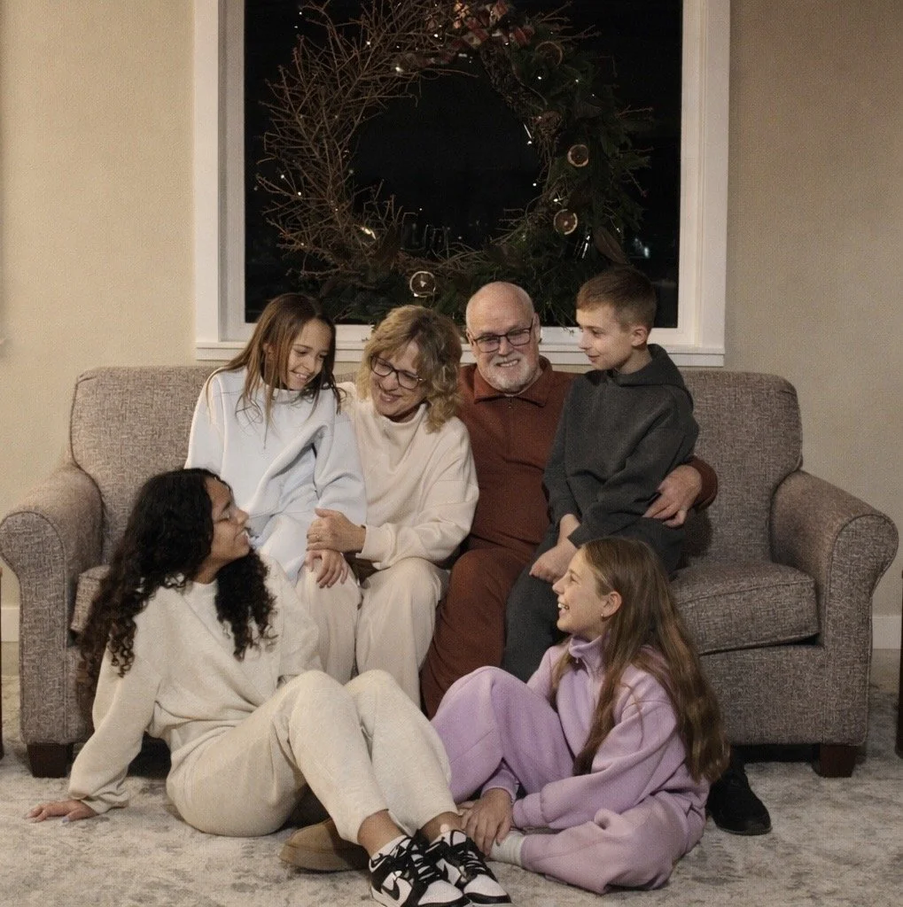 Family gathered on couch and floor around Christmas time, smiling and laughing. There is a large Christmas wreath on the window behind them.