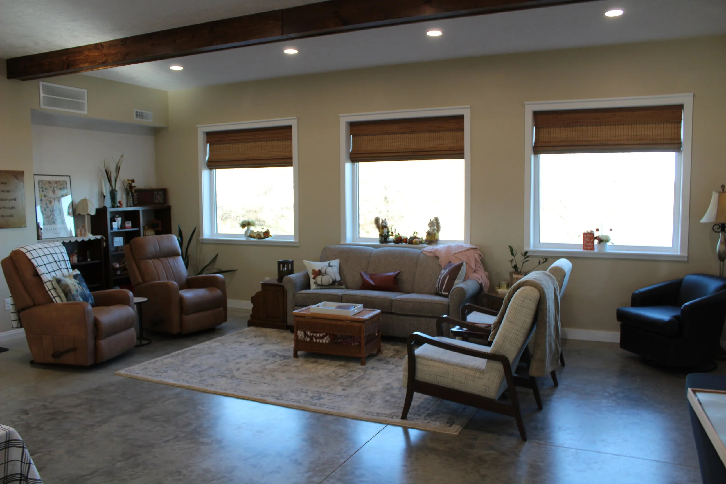 Living room with beige walls, three large windows with brown blinds, a wooden beam ceiling, and a mix of chairs and sofas. There are decorative pillows, a rug, and various decorative items on the shelves and tables.