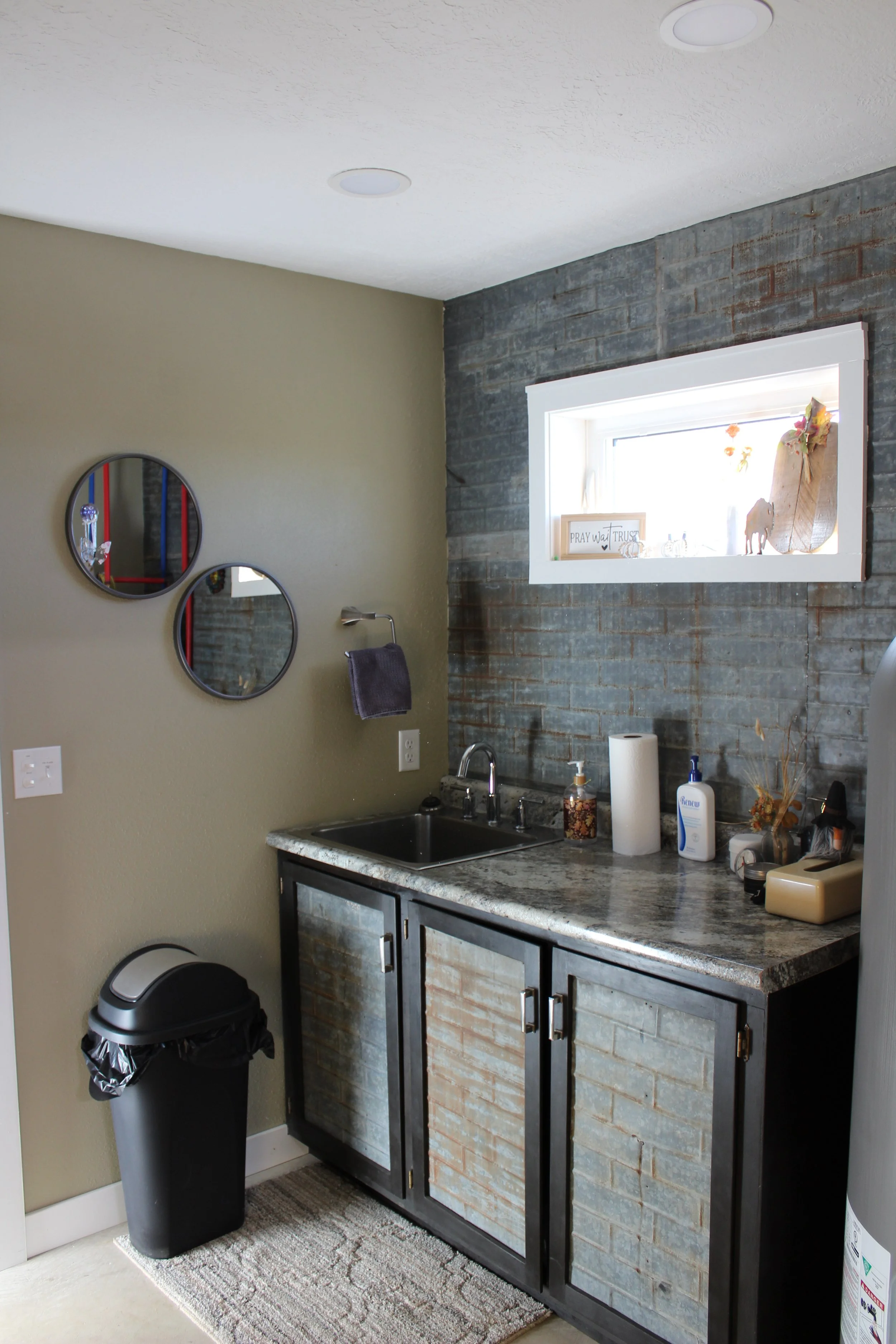 A small industrial-style kitchen area with a brick-look cabinet under a granite countertop, a small window with decorations, a sink, various bottles and supplies on the counter, and a trash can nearby.
