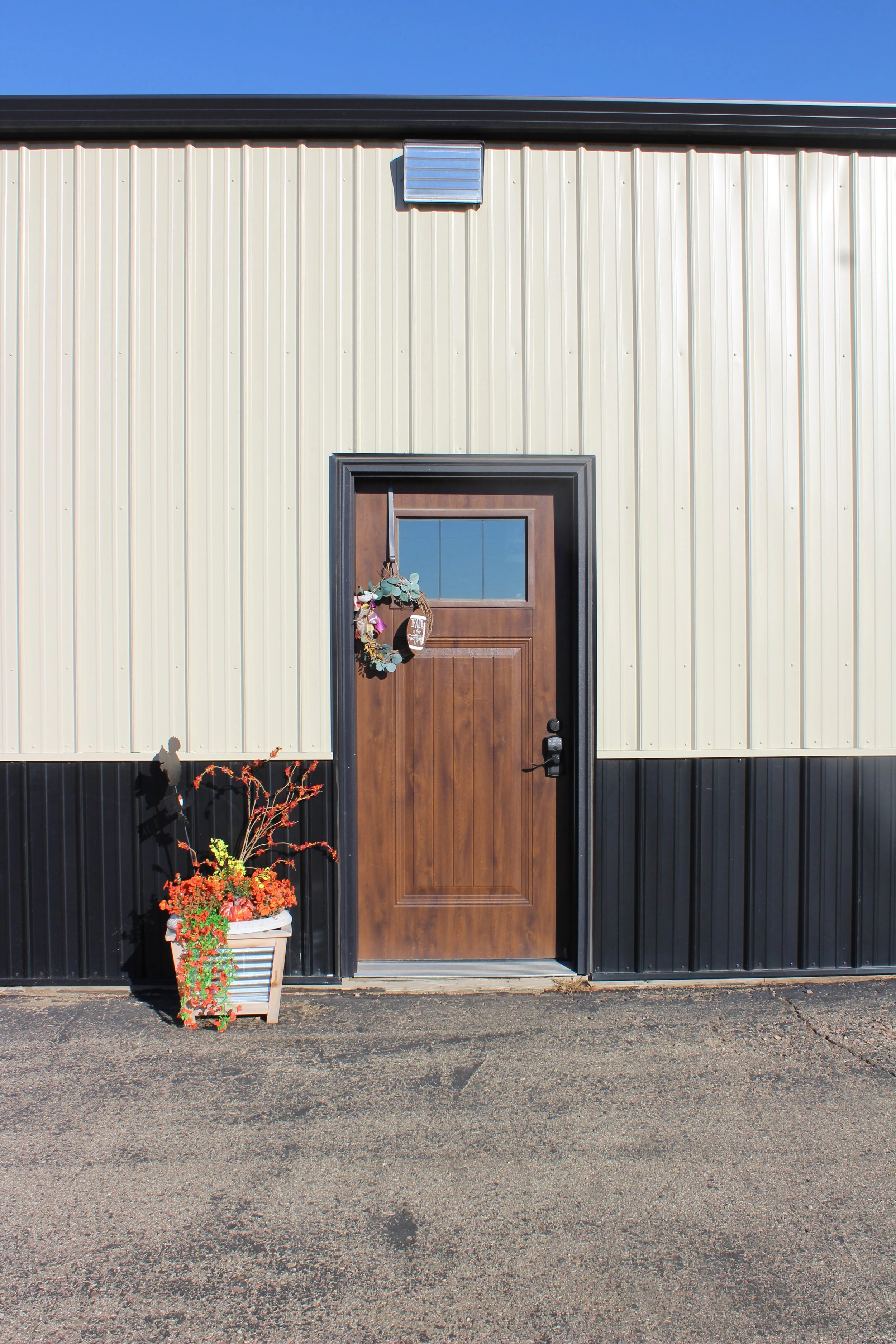 Front door of a building with beige and black siding, a small window at the top of the door, a wreath hanging on the door, and a flower pot with orange and red flowers to the left of the door.