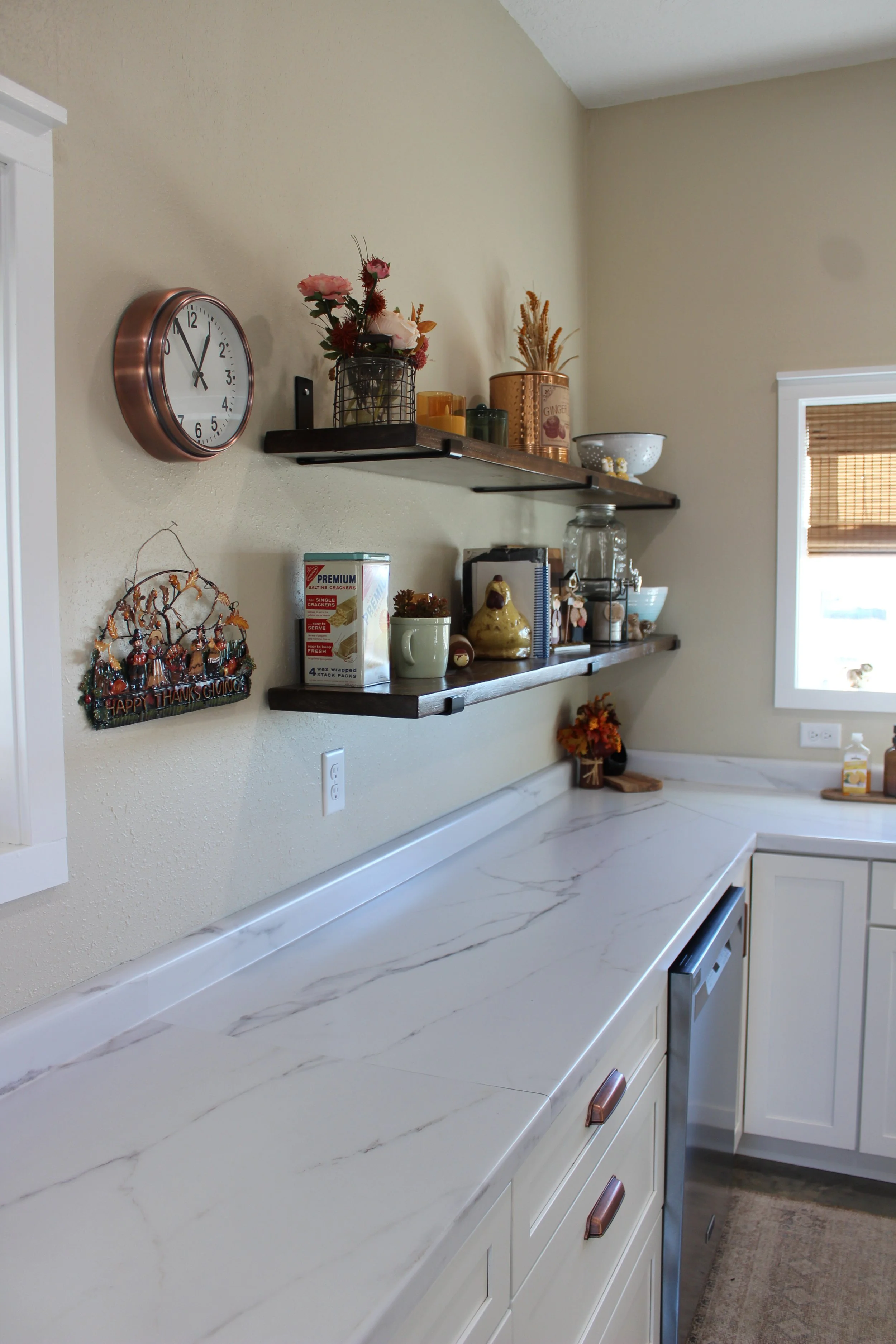 Kitchen corner with white marble countertop, white cabinets, a window with bamboo shade, and wall-mounted dark wood shelves holding decorative items and kitchen supplies.