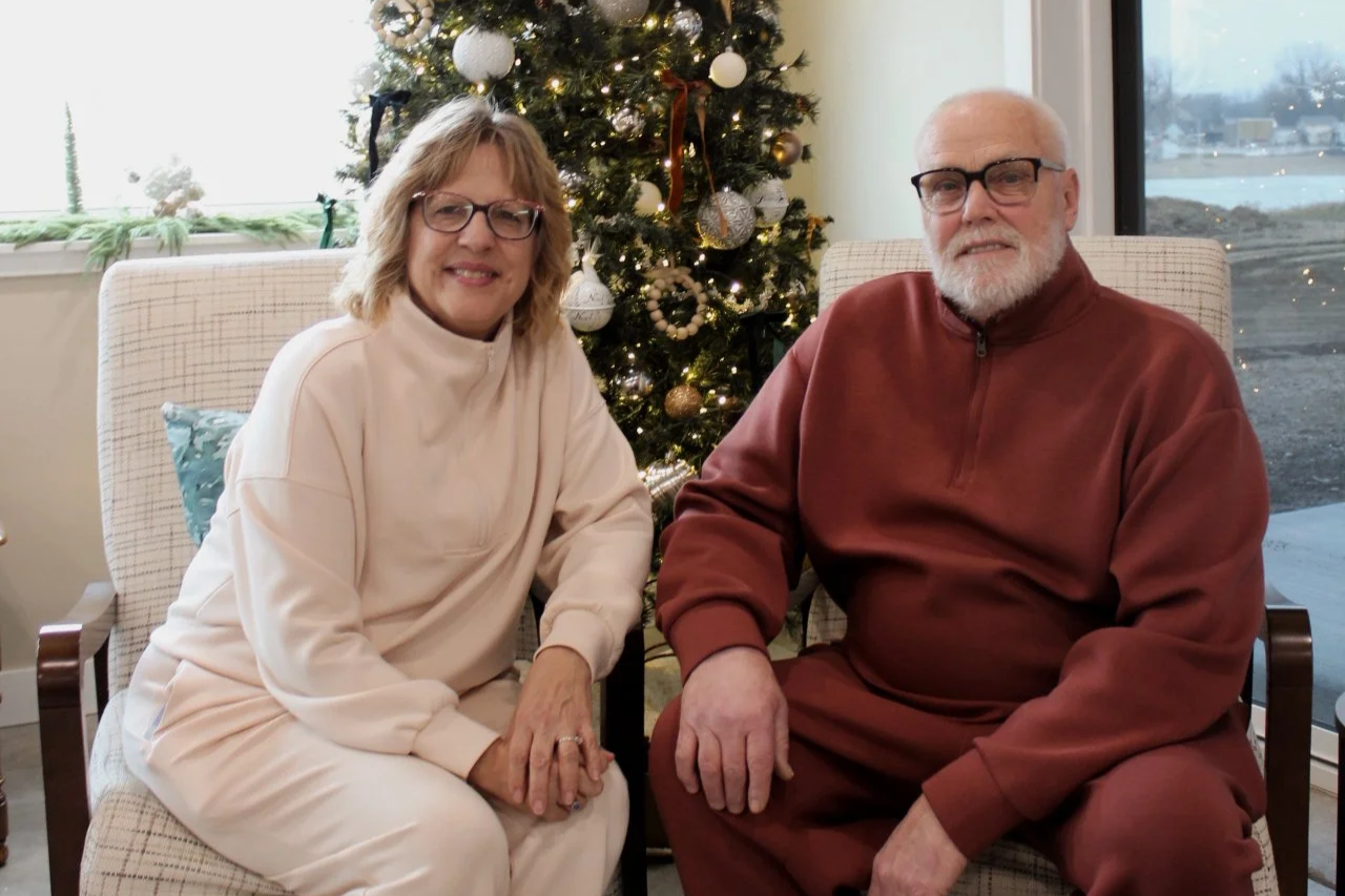 A smiling woman with glasses and blonde hair sitting next to an elderly man with glasses and a beard, in front of a decorated Christmas tree inside a room with a window showing an outdoor winter scene.