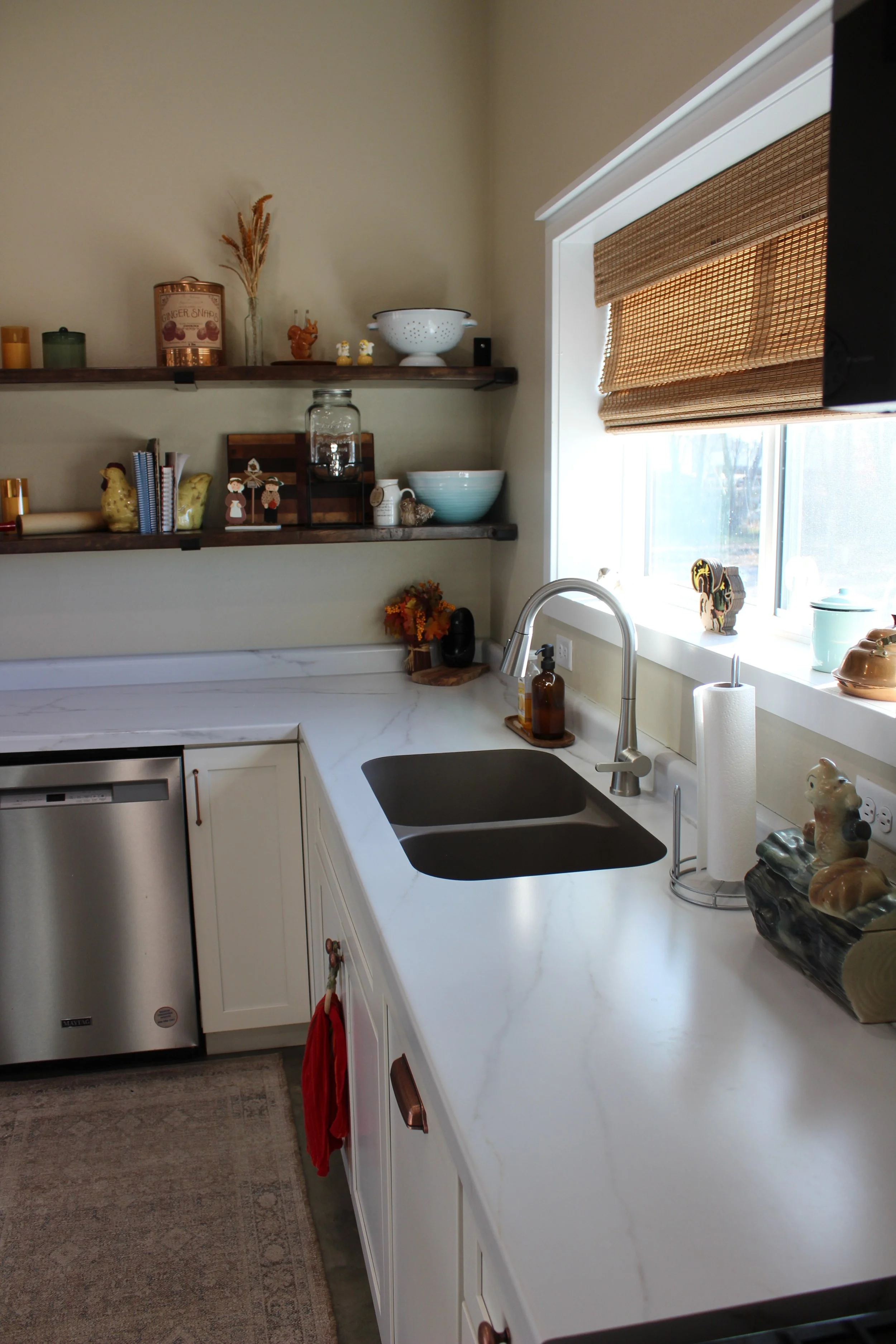 Kitchen with white countertop, black sink, window with bamboo shade, open wooden shelves with decorative items, coffee paper towel holder, and small dishwasher.