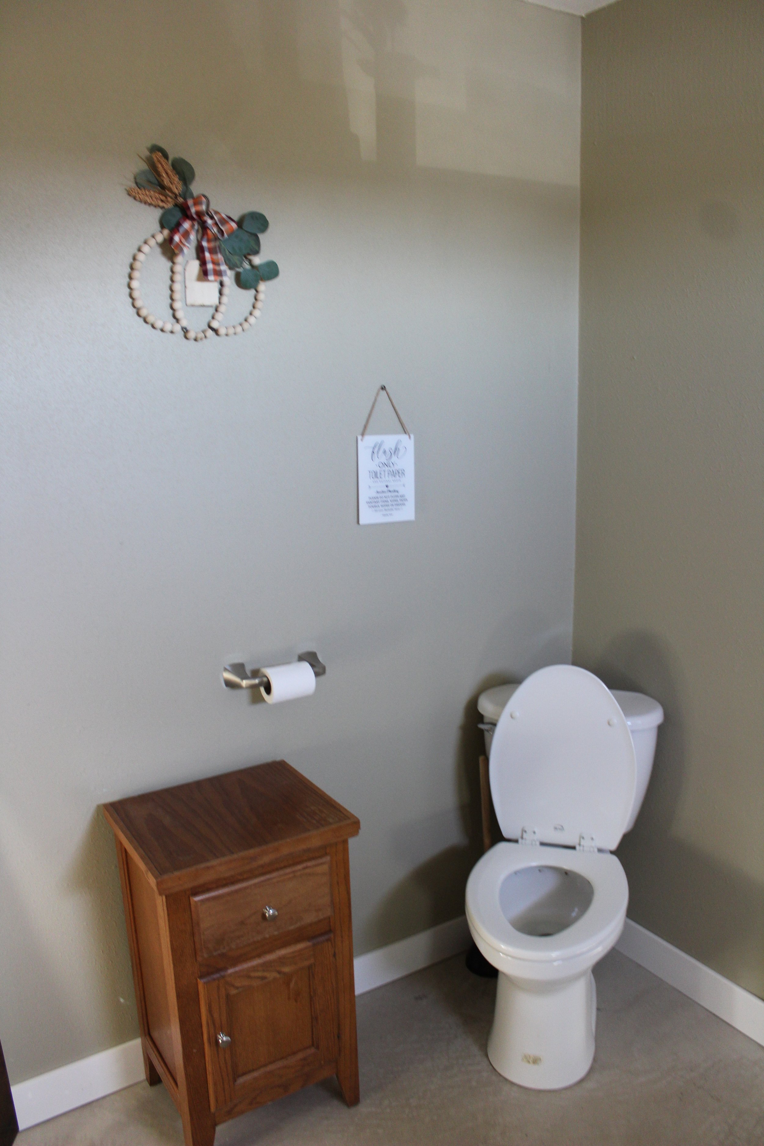 A bathroom with a toilet, a small wooden cabinet, a toilet paper holder with tissue, a decorative wall hanging, and a sign on the wall.