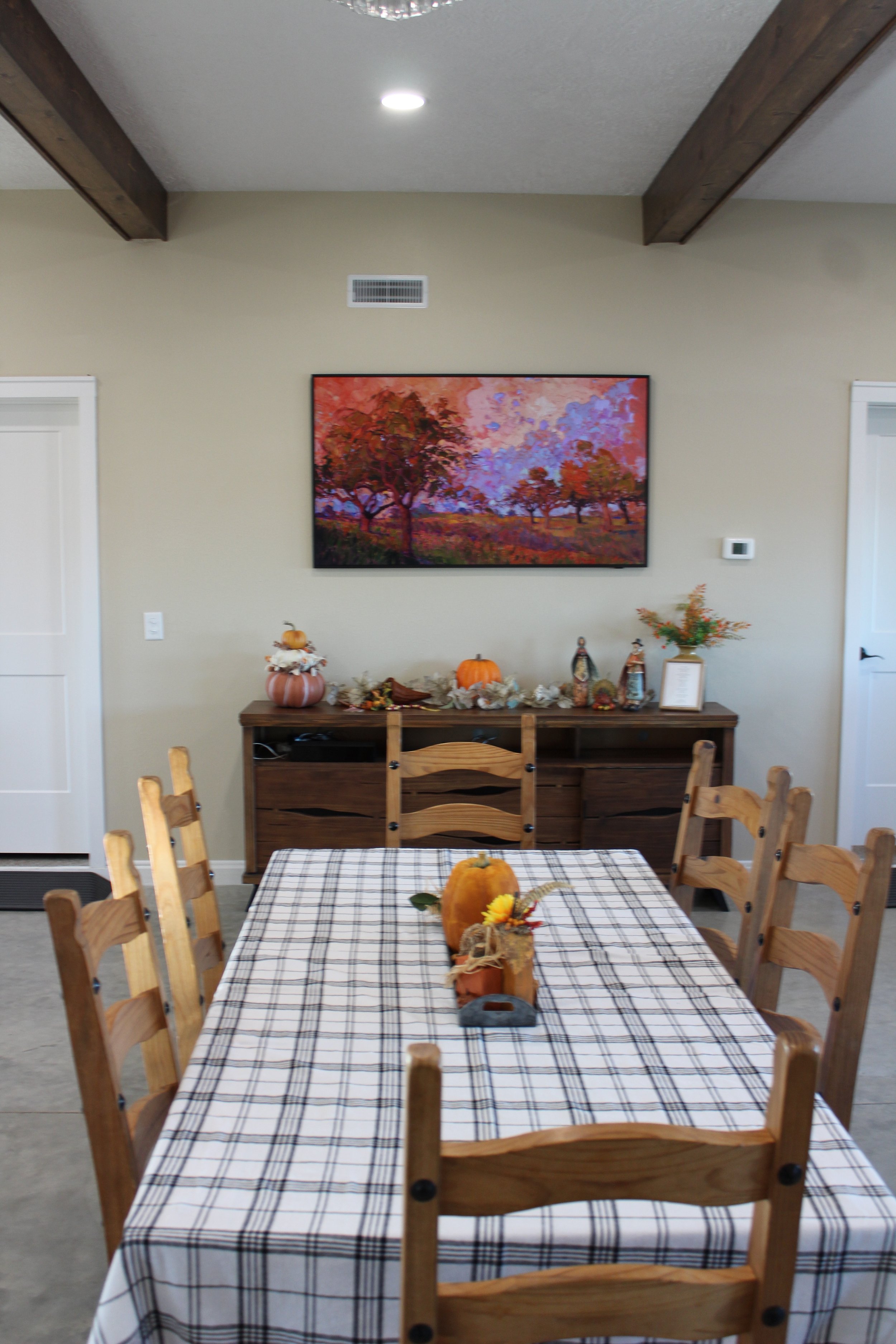 A dining room with a wooden table covered with a checkered tablecloth and decorated for fall with a pumpkin, sunflower, and small scarecrow. Six wooden chairs surround the table. A sideboard with fall decorations, including a pumpkin, scarecrows, and