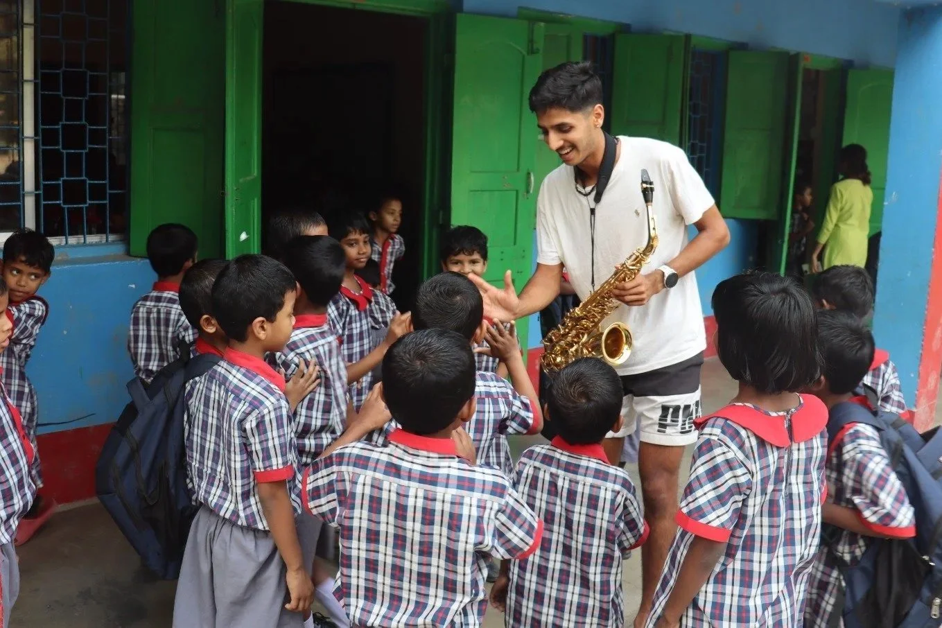 Om Satija with a saxophone smiling and reaching out to a group of young schoolchildren in uniform outside a colorful school building at the Udayan Kolkata campus.