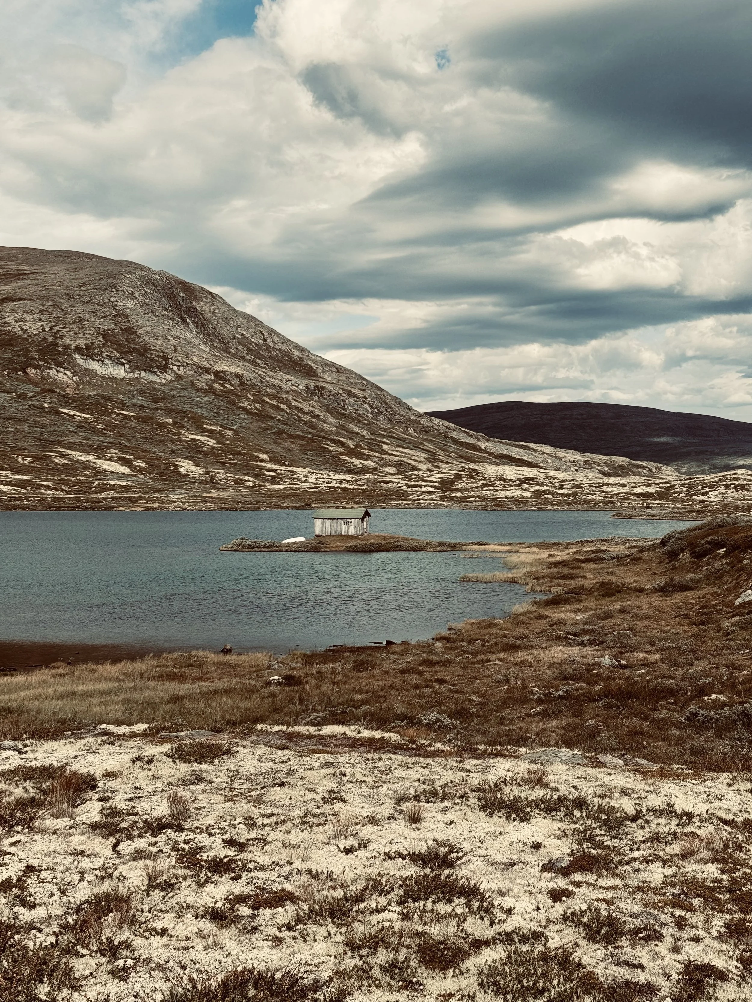 Landschaft mit einem See, umgeben von Bergen und bewölkten Himmel, und einer kleinen Hütte am Ufer des Sees.