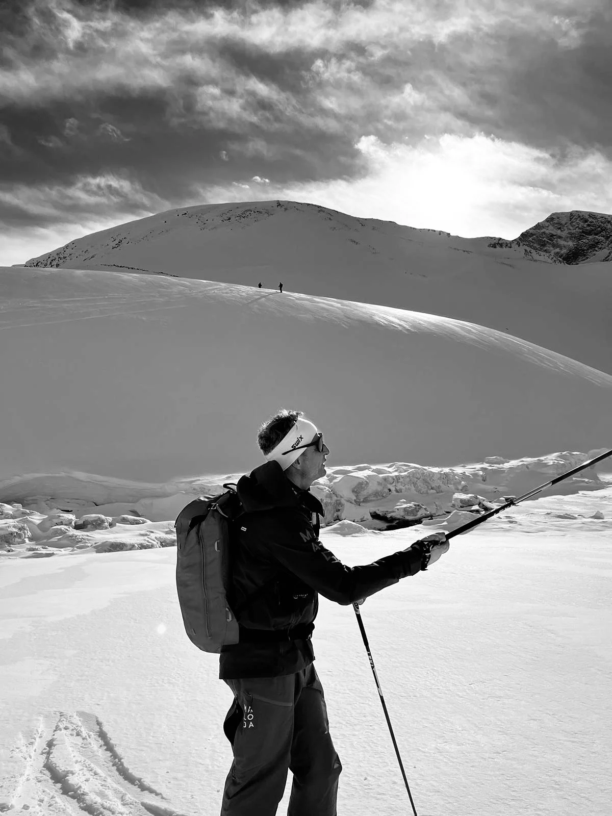 Ein Skifahrer mit Rucksack im Schnee, in der Umgebung hoher Berge und bewölktem Himmel, im Schwarz-Weiß-Format.