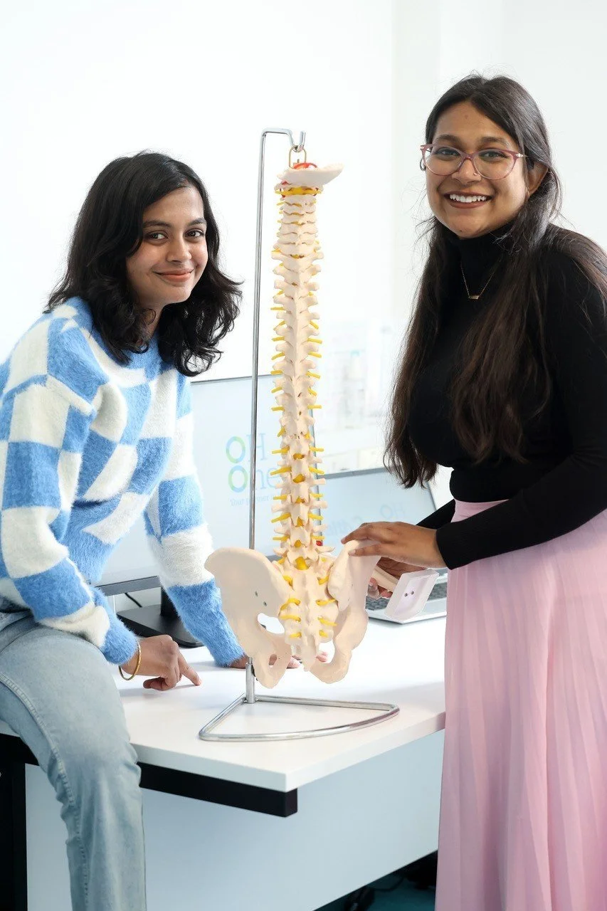 Two women smiling at a table with a model of a human spine and pelvis.
