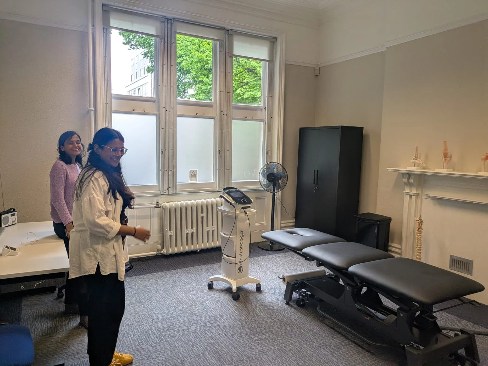 A physiotherapy or chiropractic room with two women standing and smiling, one in white and the other in pink, near a treatment table, with medical equipment including a technician device on wheels, a fan, a black cabinet, and a model of a human spine on the fireplace mantel.