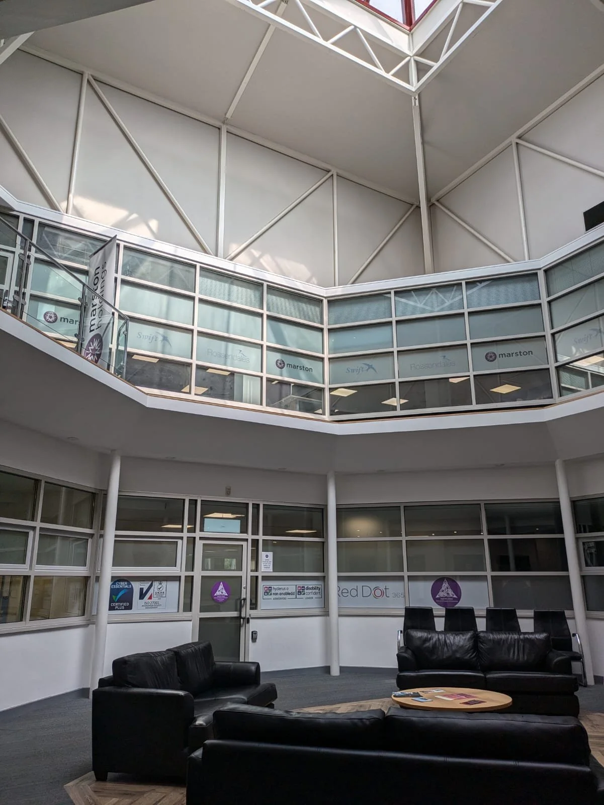 Photograph of the internal lobby of Triangle Building Centre where OH One Physiotherapy clinic is based alongside Case UK. This is a large modern looking octagon shaped glass atrium, with black leather sofas surrounding a coffee table, waiting area.