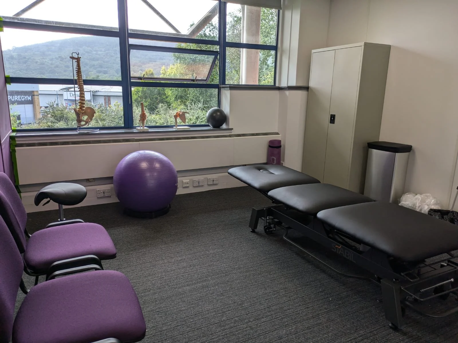 Photograph of the OH One Physiotherapy treatment room within Triangle Building Centre (Merthyr Tydfil). The room features a black physiotherapy treatment bed, purple chairs and exercise rehab equipment.