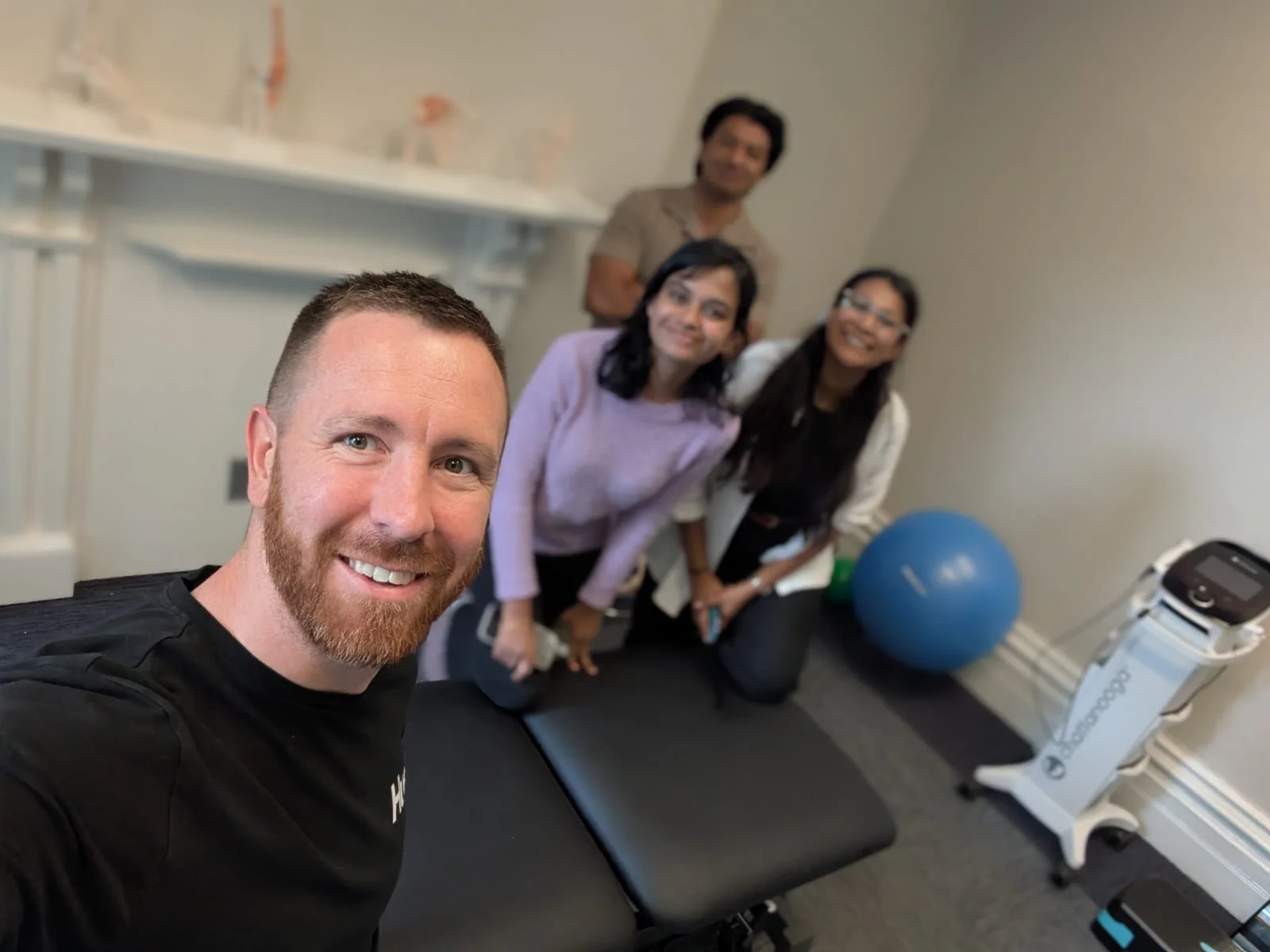 Group of four people in a medical or fitness setting, smiling for a selfie, with exercise equipment including a large blue exercise ball and a medical device visible in the background.