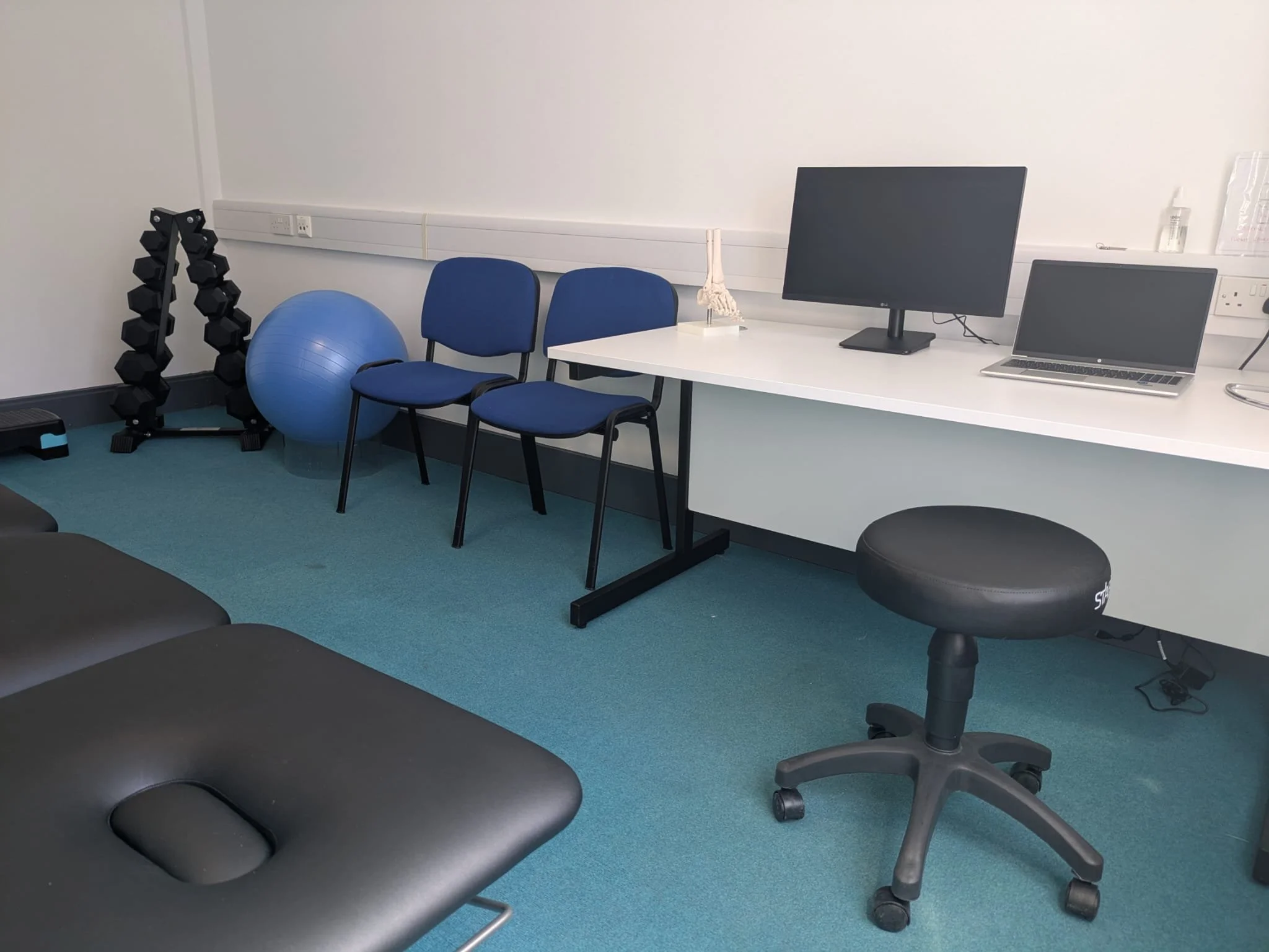Medical or physiotherapy examination room with a massage table, a weight stand, a blue exercise ball, two blue chairs, a desk with a monitor, a laptop, a model of a human foot, and a black technician stool.