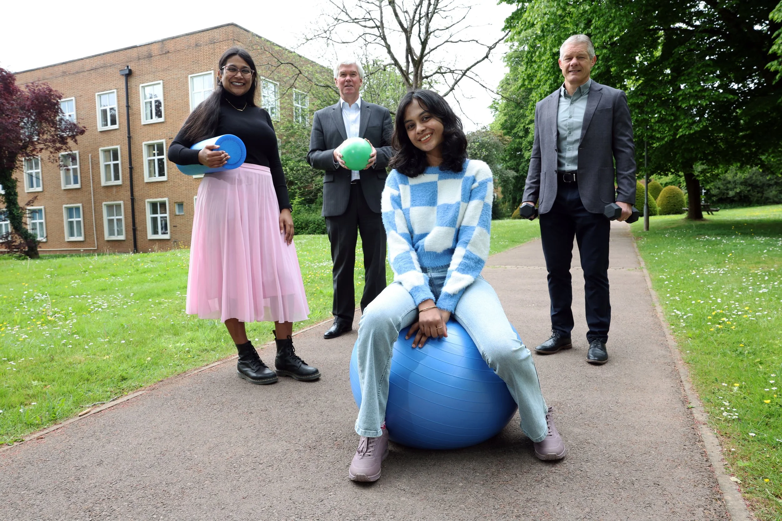 Group of four adults and one young woman sitting on an exercise ball outdoors in a park, smiling at the camera, with buildings and trees in the background.