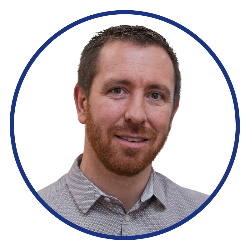 A portrait of a smiling man with short brown hair and a beard, wearing a light gray collared shirt, against a white background with a blue circular border.
