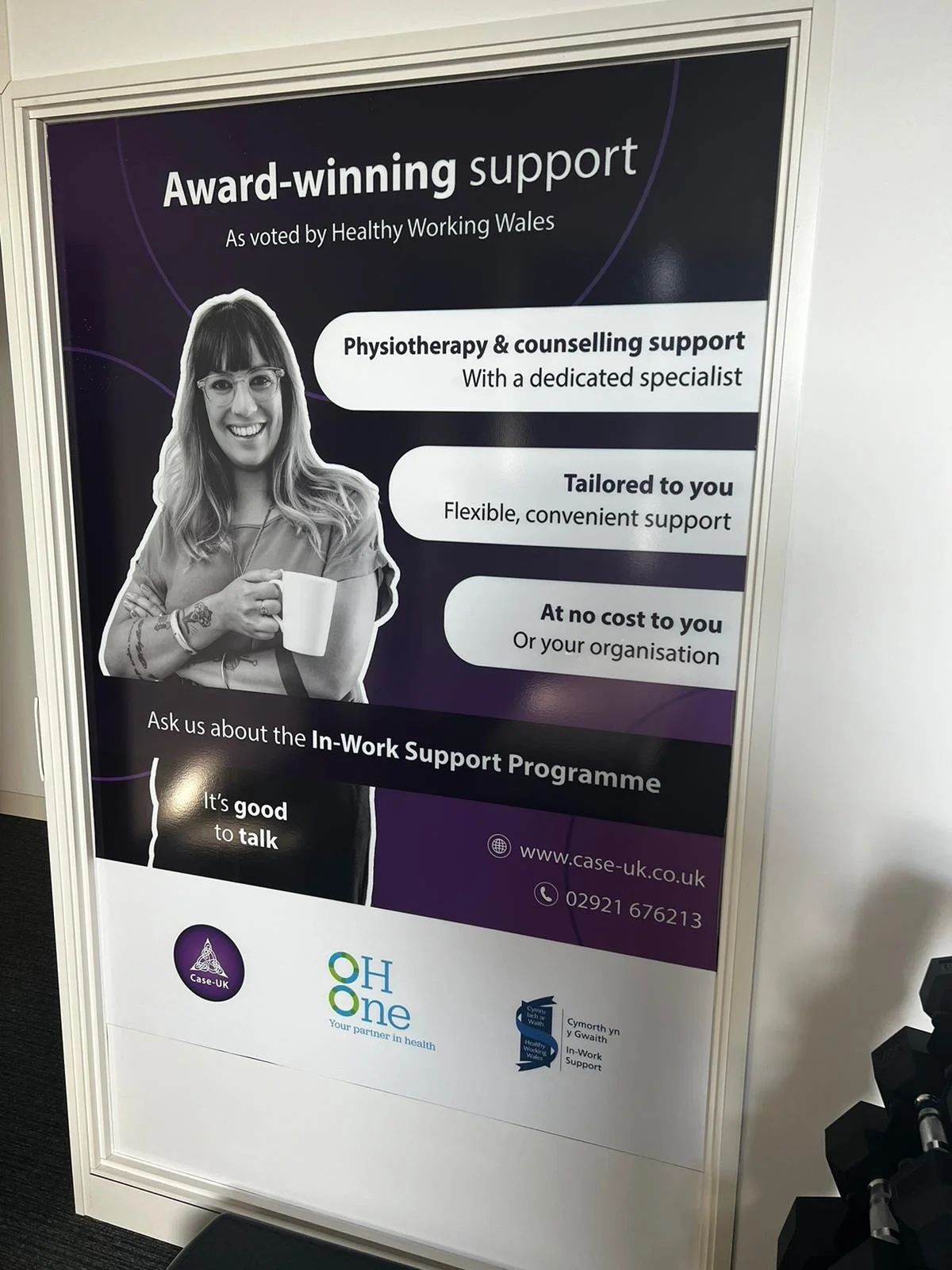 A photograph of a large window vinyl providing patients with information about the Wales Government In-Work Support Service. This explains that in partnership with Case UK, OH One offer physiotherapy and counselling to employees in Wales.