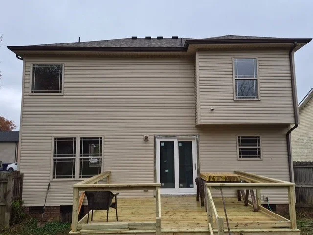 The back of a beige two-story house under construction with a wooden deck, patio furniture, and a sliding glass door.