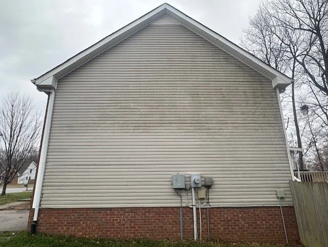 The side of a house with beige vinyl siding, a brick foundation, electrical meters, and pipes, with a yard and trees in the background.