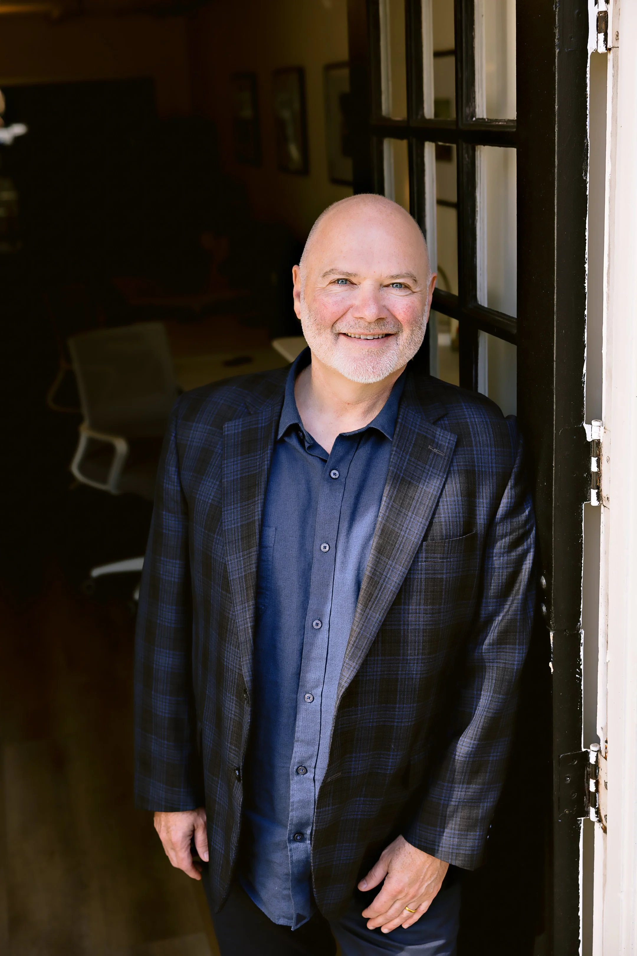Gary is smiling, wearing a dark plaid blazer and a dark blue shirt, standing near a window with black panes, inside a room with framed pictures on the wall.
