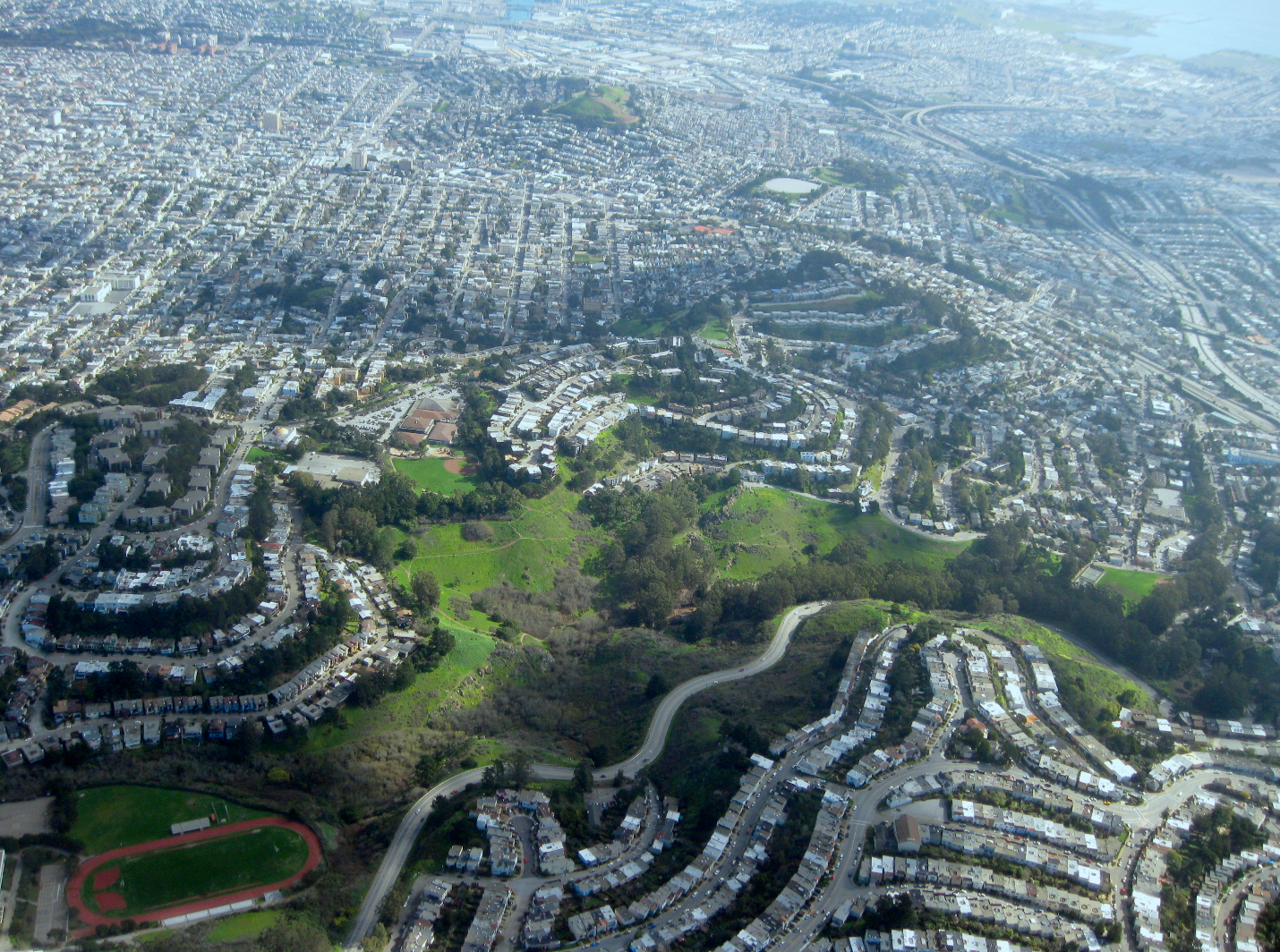 An aerial view of a densely populated urban area with residential neighborhoods, roads, and green parks, including a sports track.