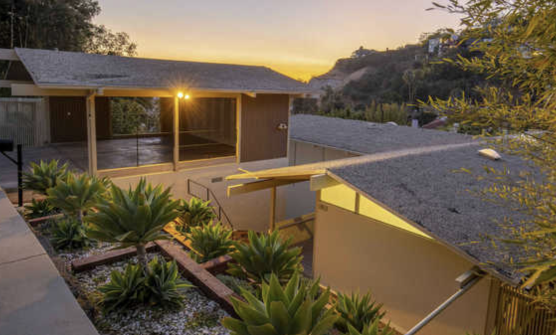View of a modern house's backyard during sunset, with a garden of succulents, neighboring houses, and rolling hills in the background.