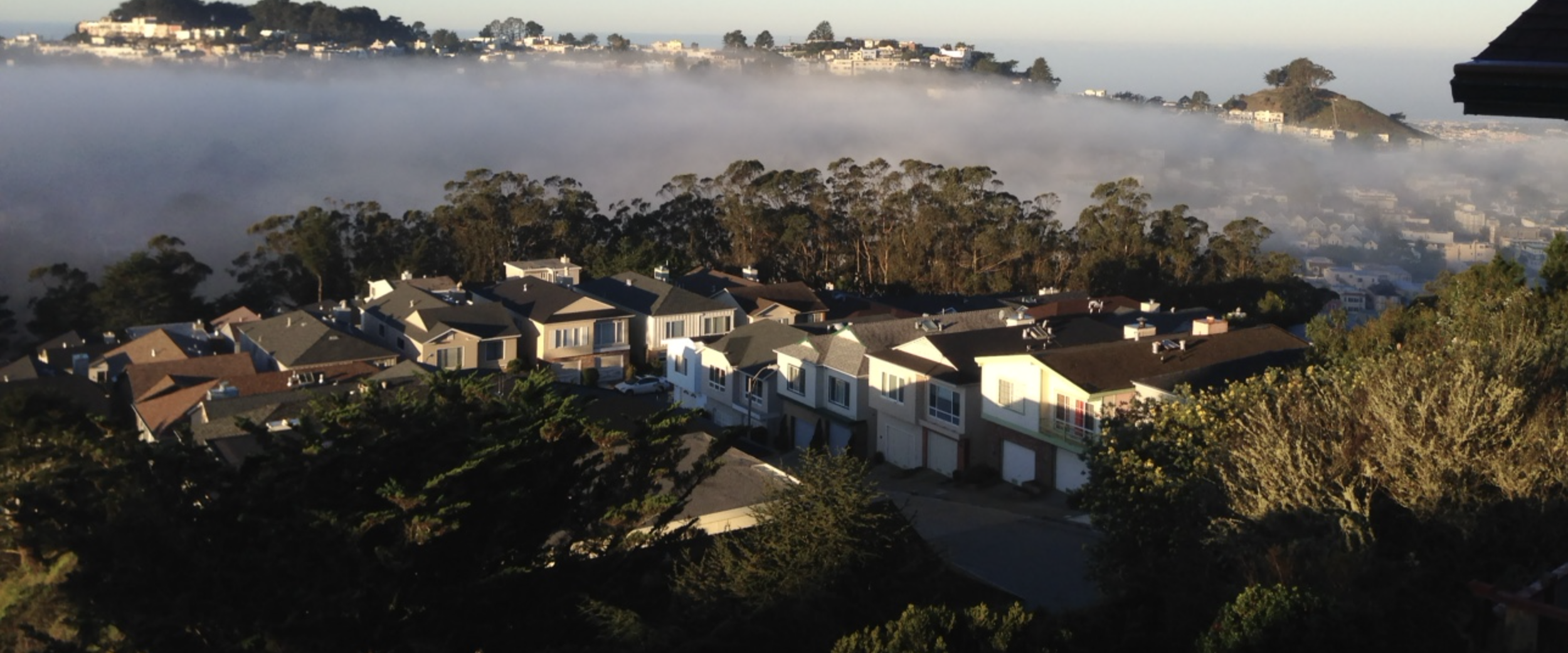 Residential neighborhood with modern houses, trees, and fog in the background overlooking hilly terrain at sunrise or sunset.