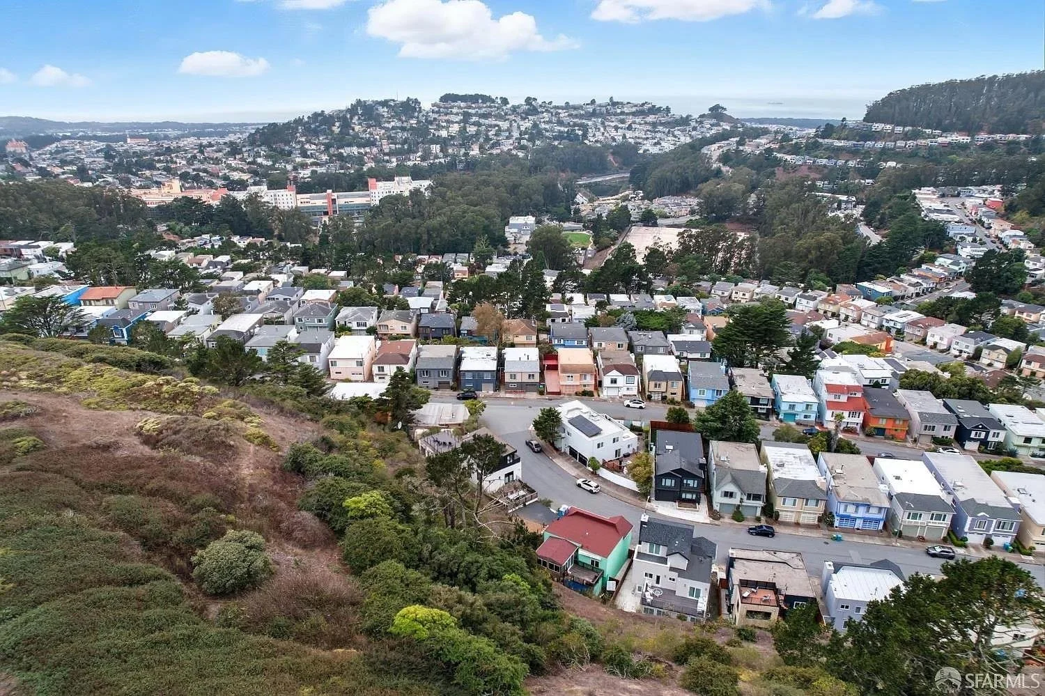 Aerial view of a hillside neighborhood with colorful houses and winding roads, overlooking a cityscape with numerous buildings and greenery under a partly cloudy sky.