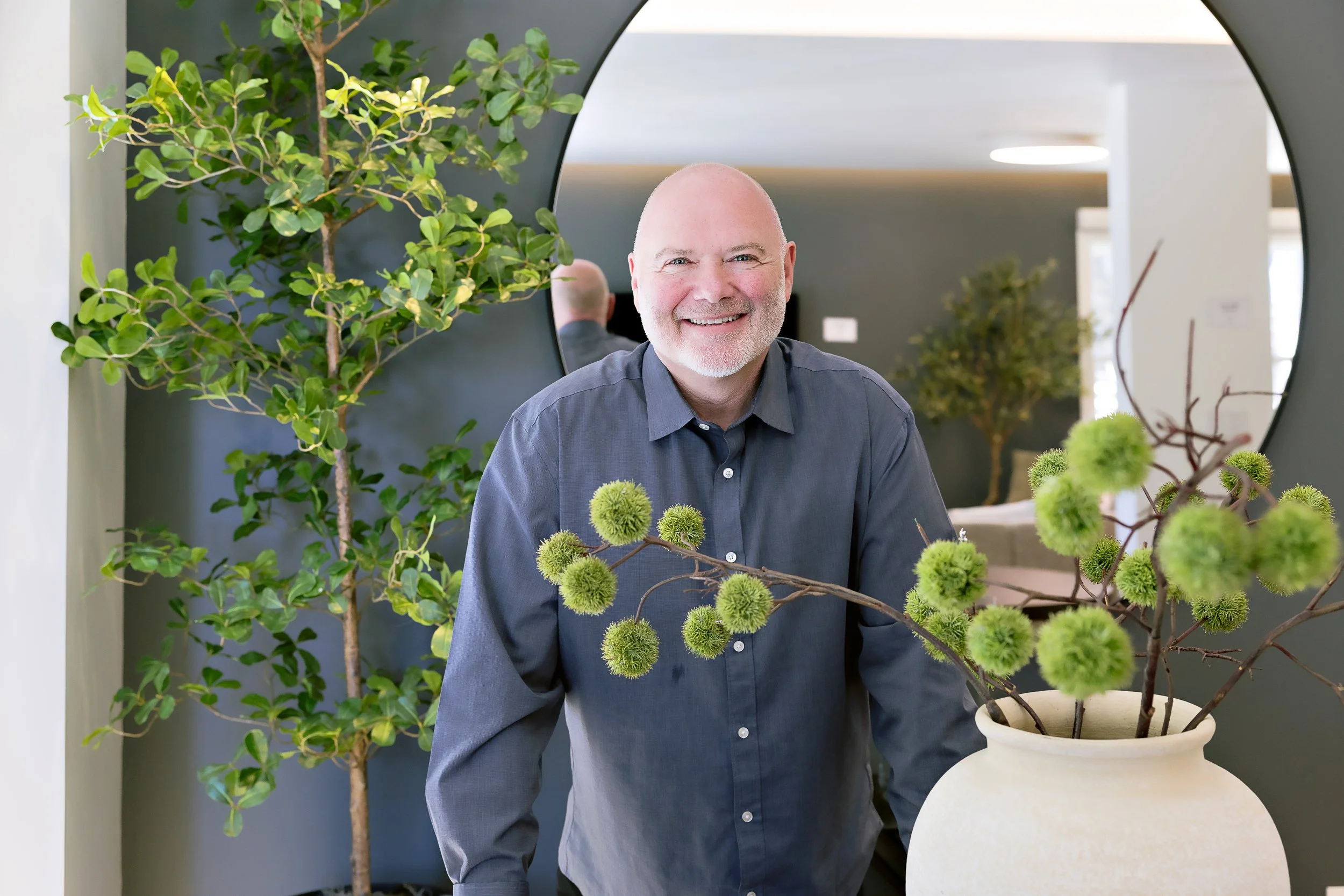 Gary is smiling, wearing a dark gray shirt, standing next to a large potted plant with green spherical flowers, in a modern indoor setting with green leafy plants and a mirror in the background.