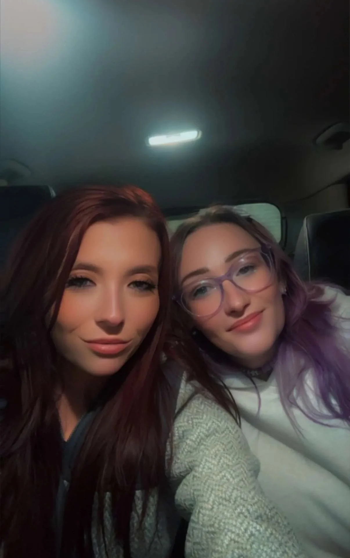 Two women with long hair, one with reddish-brown hair and the other with purple hair and glasses, sitting in a car at night, smiling at the camera.