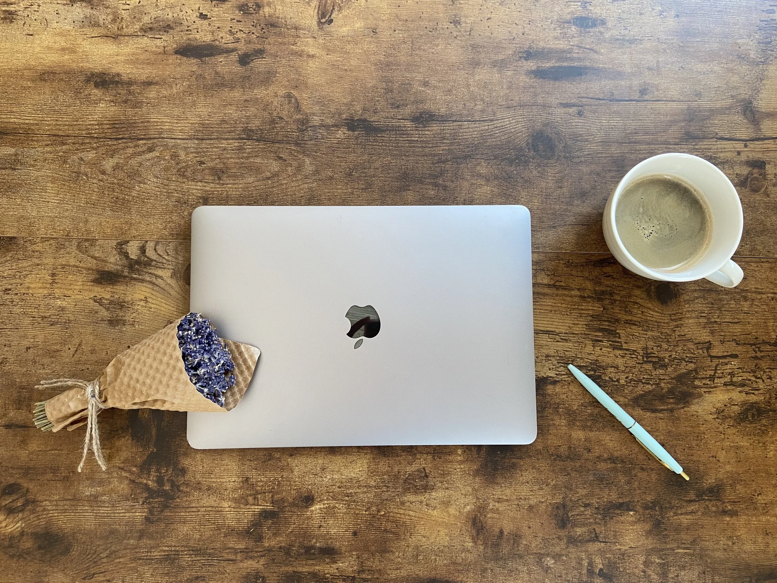 Top-down view of a wooden table with a closed silver laptop, a cup of coffee, a light blue pen, and a small bouquet of lavender flowers wrapped in brown paper and tied with string.