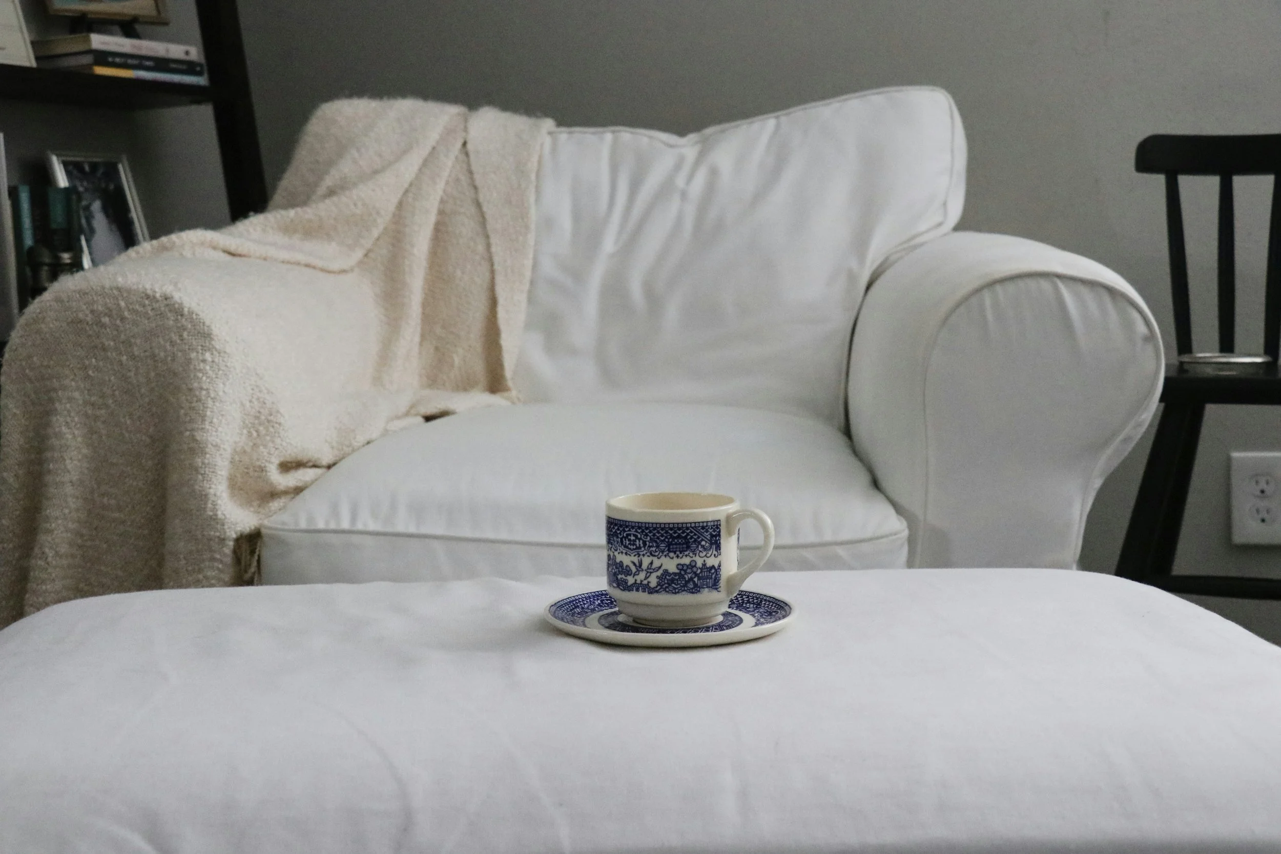 A coffee cup with a matching saucer in front of a white sofa with a beige throw blanket draped over the armrest, in a cozy living room, representing a soft place to land for clients experiencing burnout.