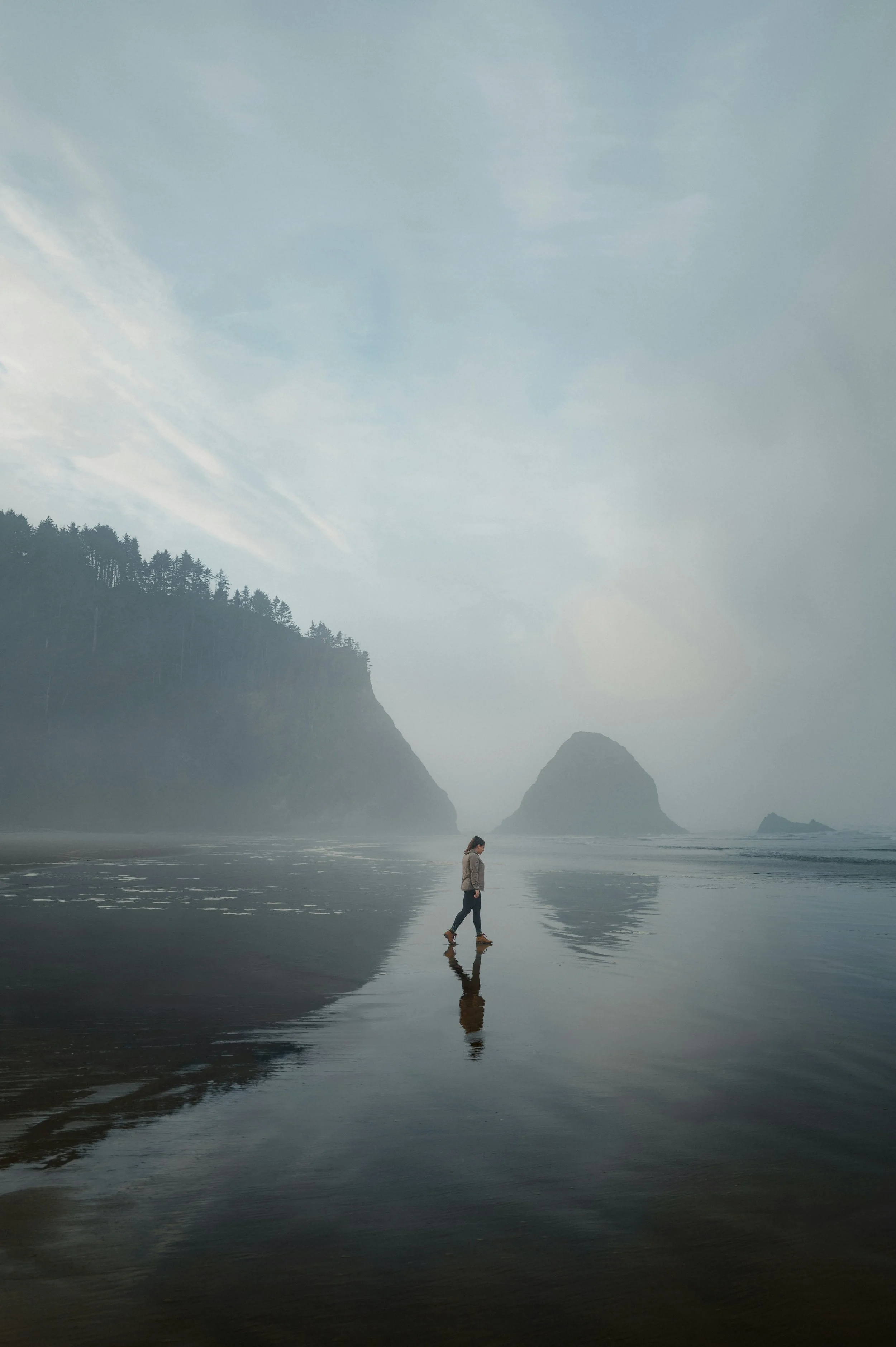 A person walking on a wet beach with misty cliffs and large rocks in the background.