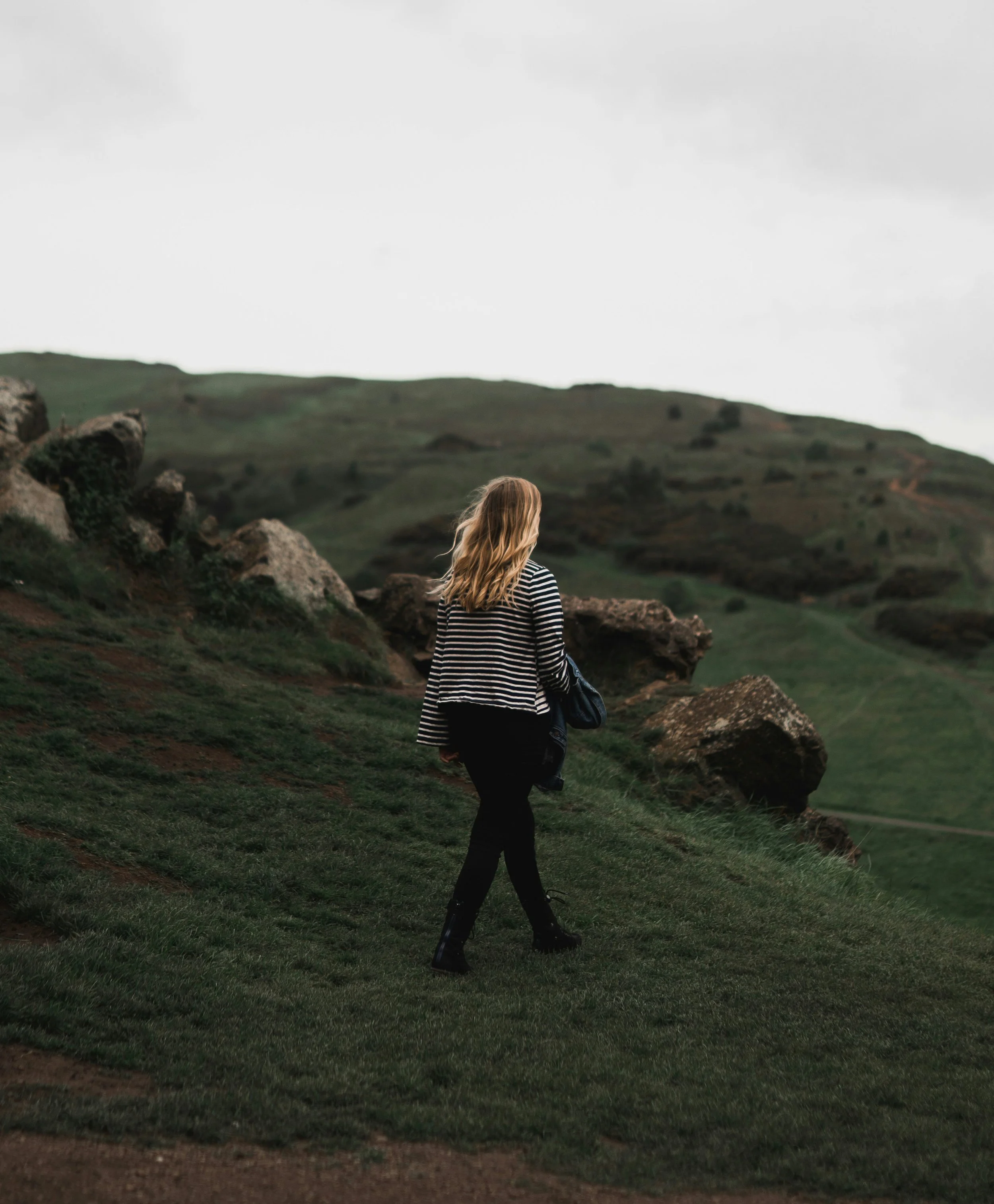 A woman with blonde hair wearing a striped black and white top, black pants, and boots walking on a grassy hillside with rocks, under a cloudy sky.