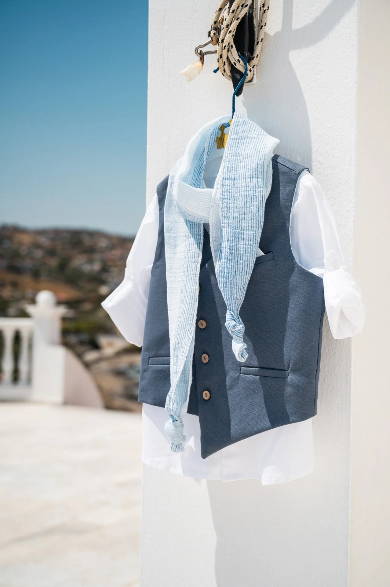 A white shirt with a blue vest and a striped towel hanging on a hook on a white wall with a clear sky background.