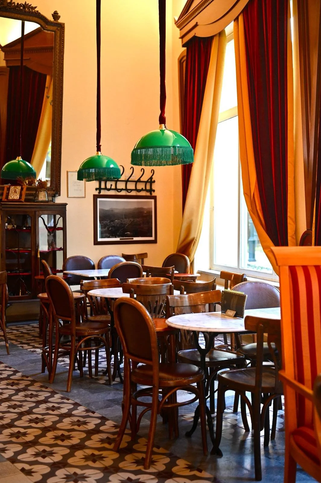 Interior view of a cozy restaurant or cafe with wooden tables and chairs, large windows with red and yellow drapes, and green pendant lamps hanging from the ceiling.