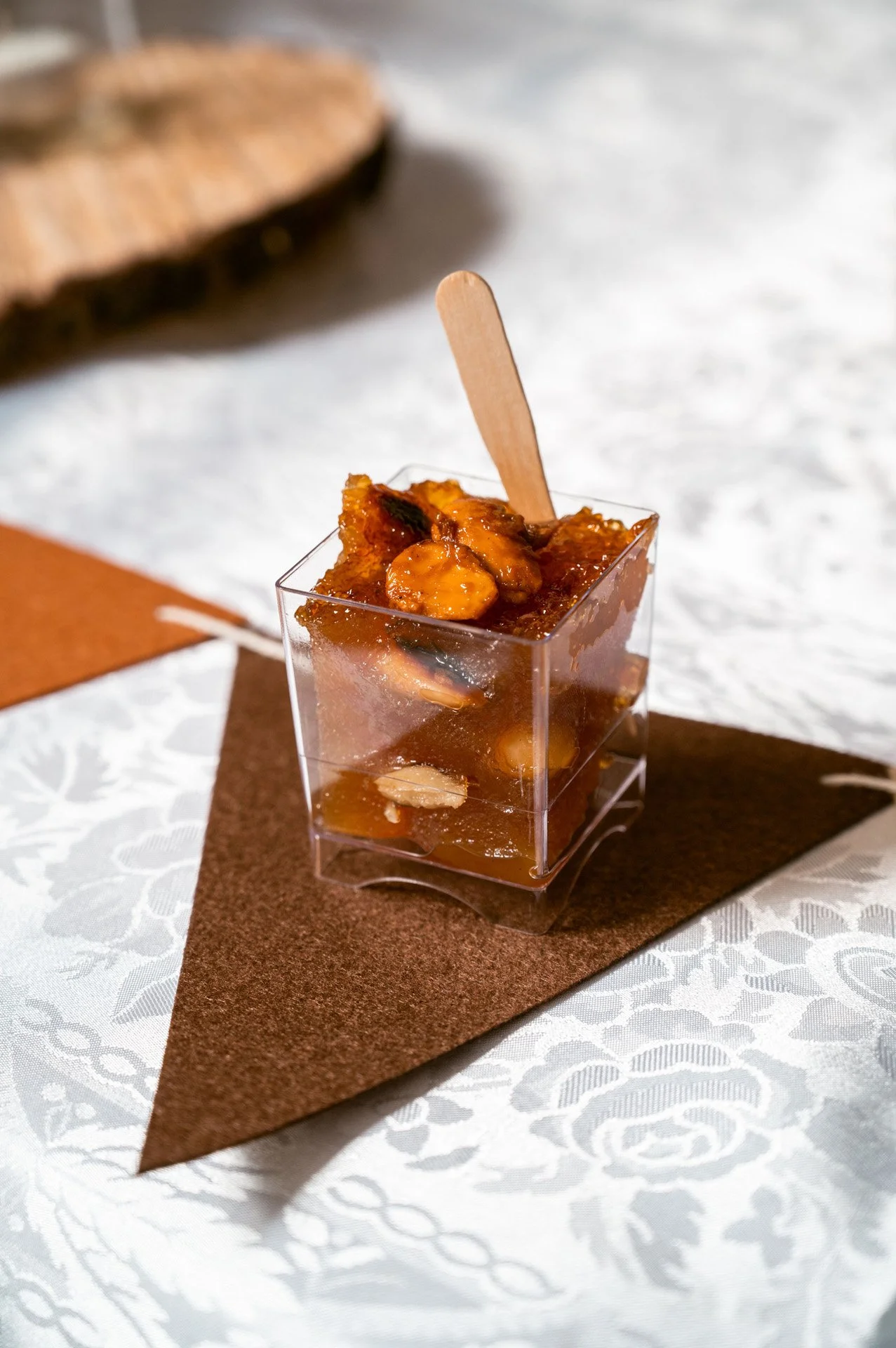 A small clear square container filled with a chunky, orange-brown chutney or relish, garnished with a small wooden stick, placed on a piece of brown paper on a white patterned tablecloth.