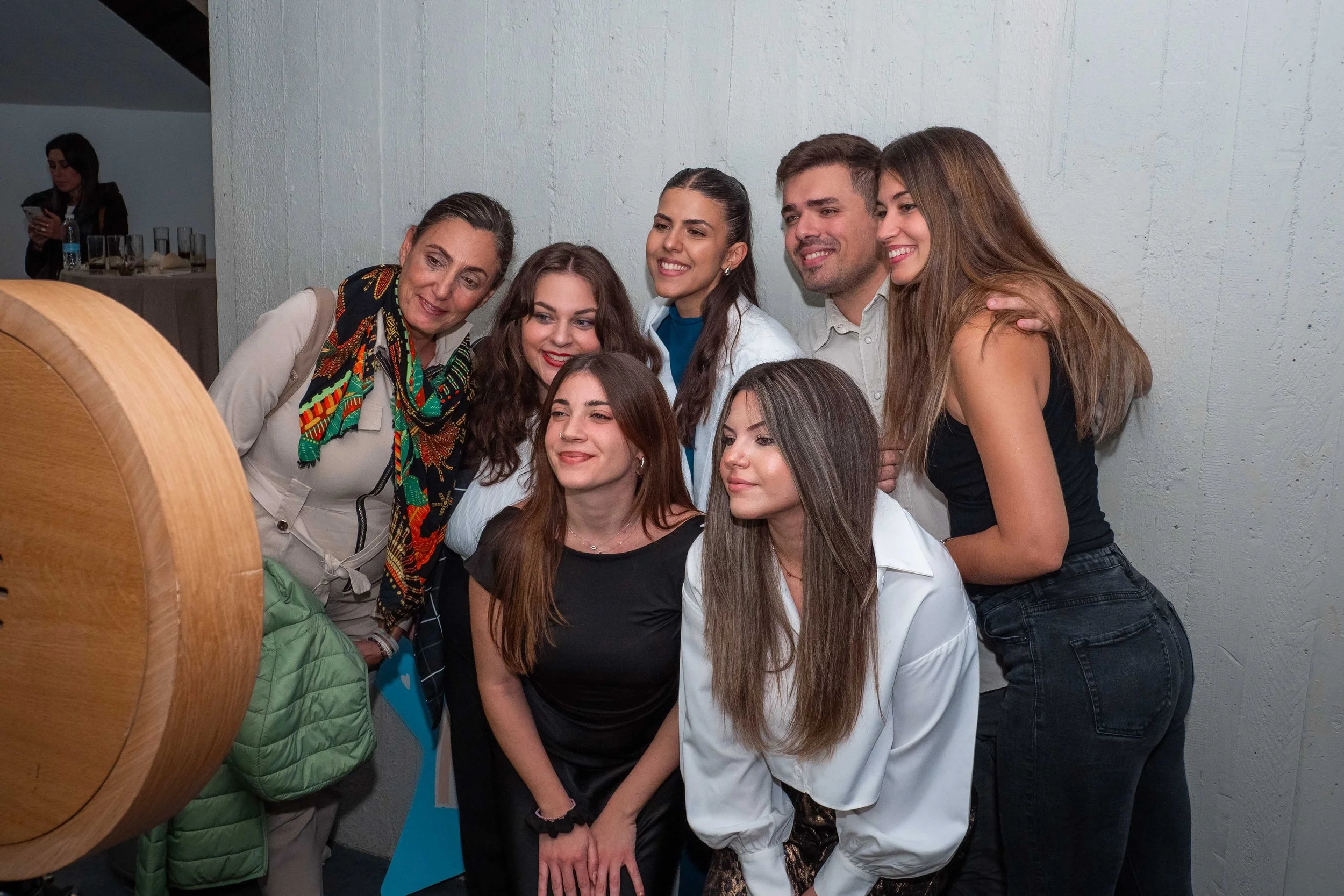 Group of seven young adults taking a photo together, smiling and posing against a white wall, with a woman in the background using a phone.