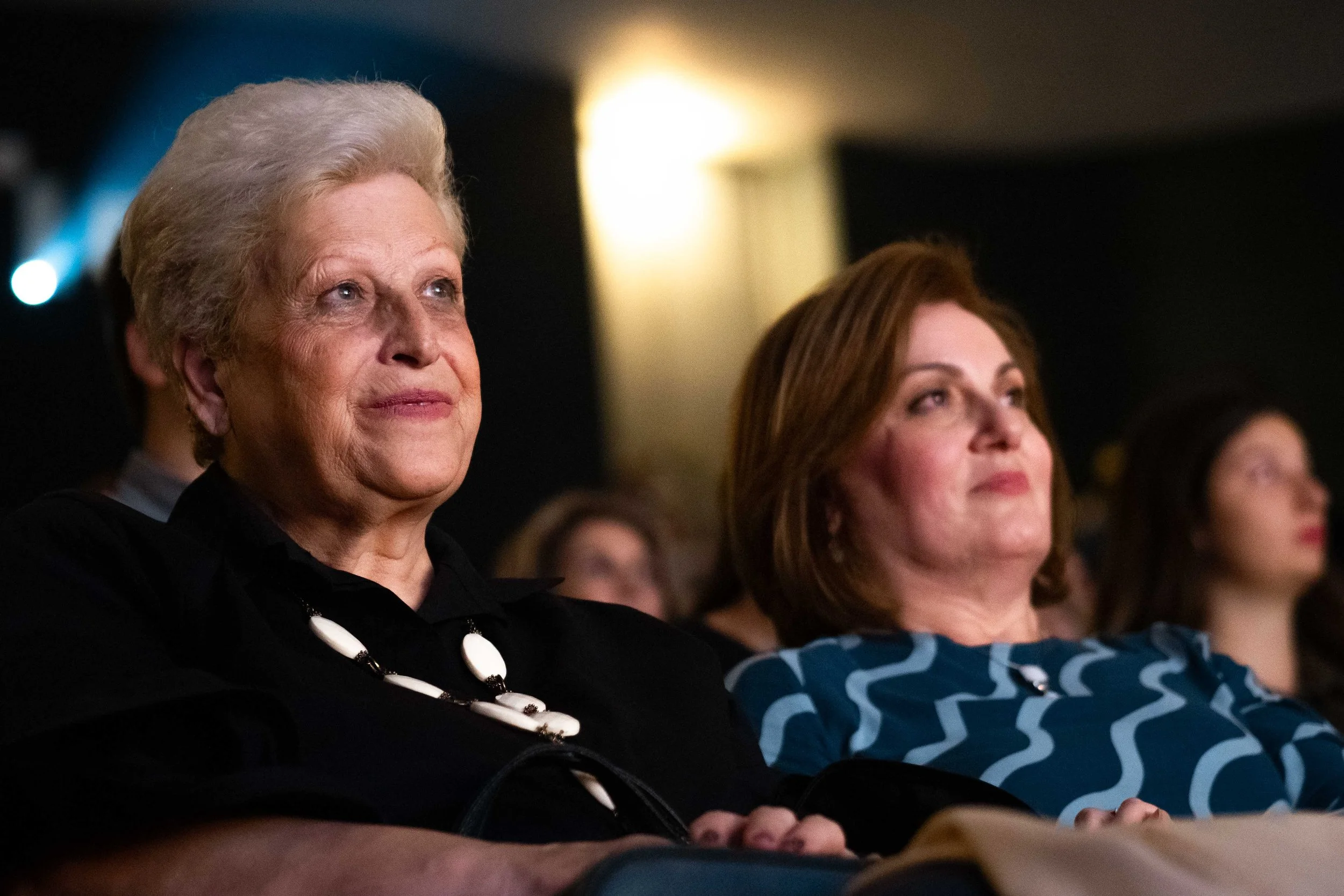 Two women seated in an audience, attentively watching a performance or presentation, with a dark background and warm lighting.