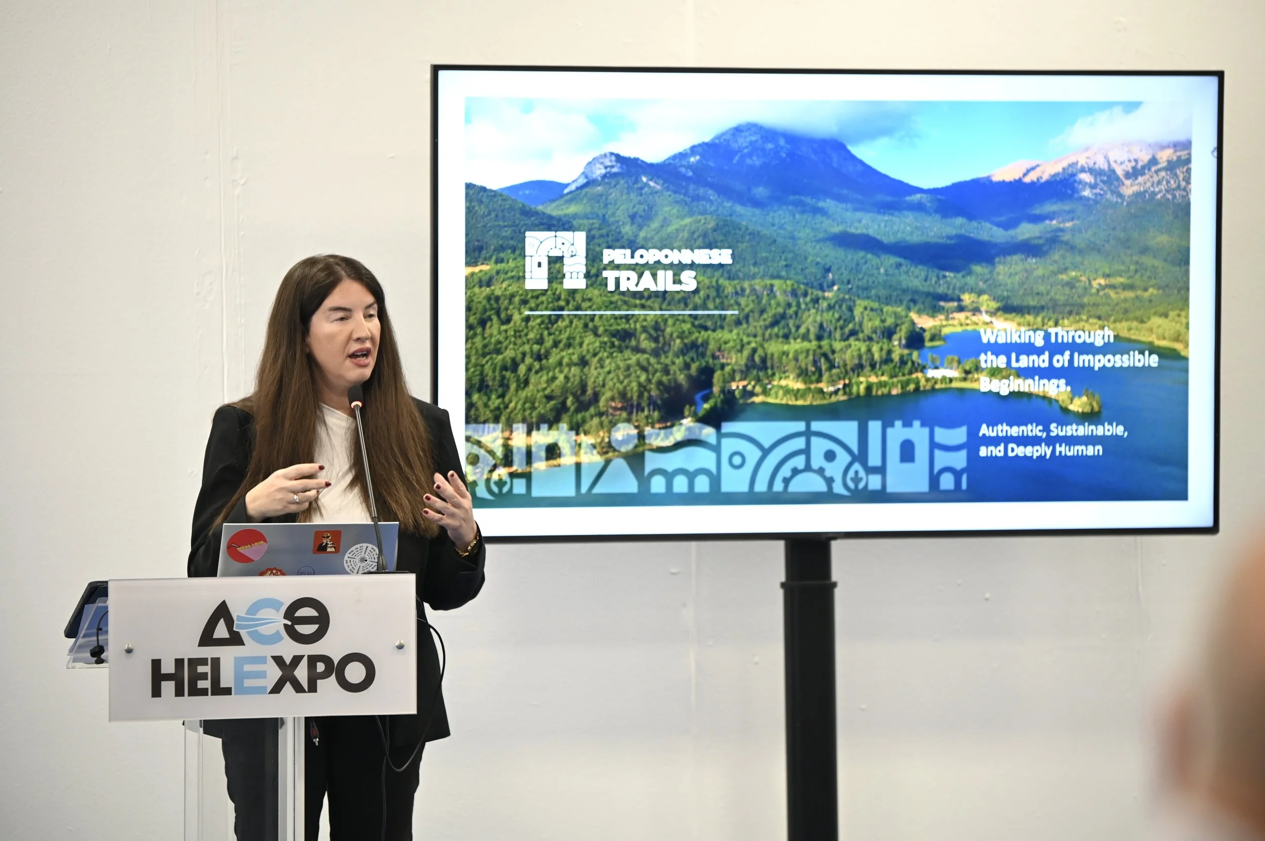 A woman giving a presentation at a conference, standing behind a podium with a HEL EXPO sign, in front of a large screen displaying a landscape of mountains and a lake with text about Peloponnese Trails.