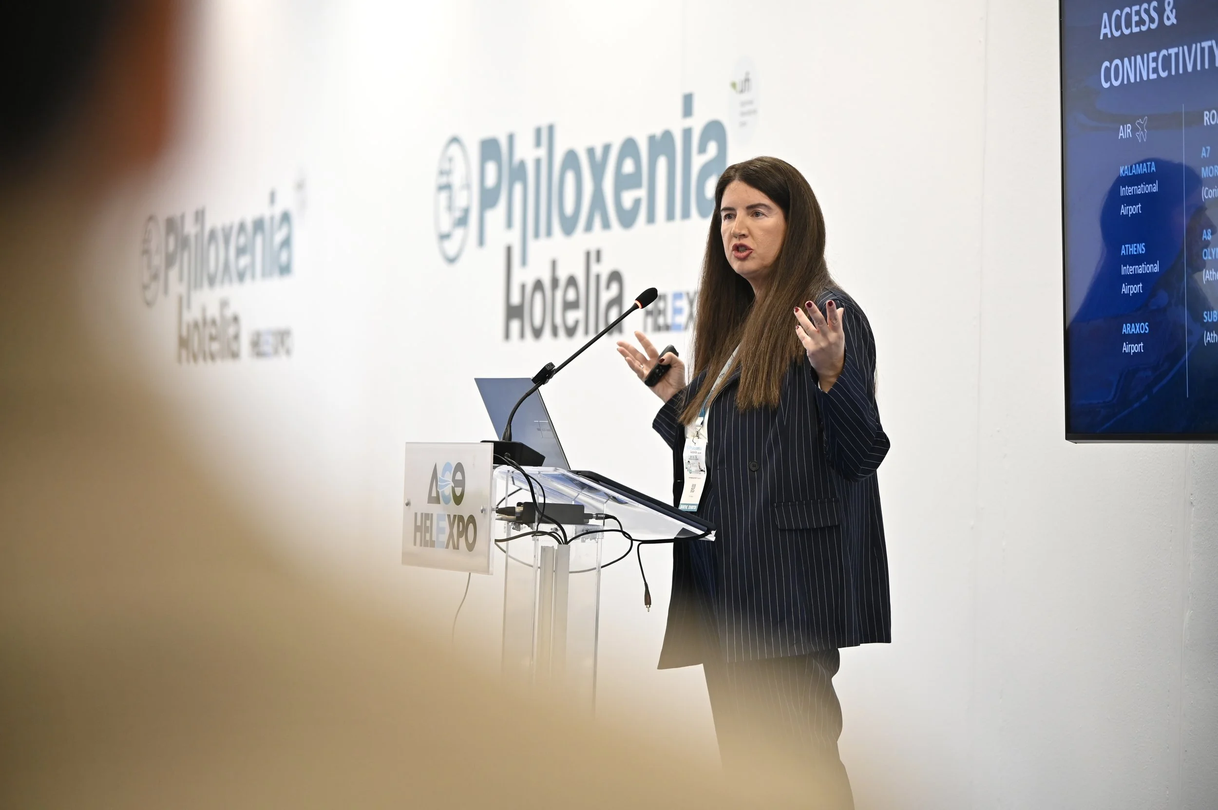 A woman with long brown hair presenting at a conference, standing behind a podium with a microphone. She is wearing a dark pinstripe blazer and gesturing with her hands. Behind her are large logos of Philoxenia Hotelia and a screen displaying confere