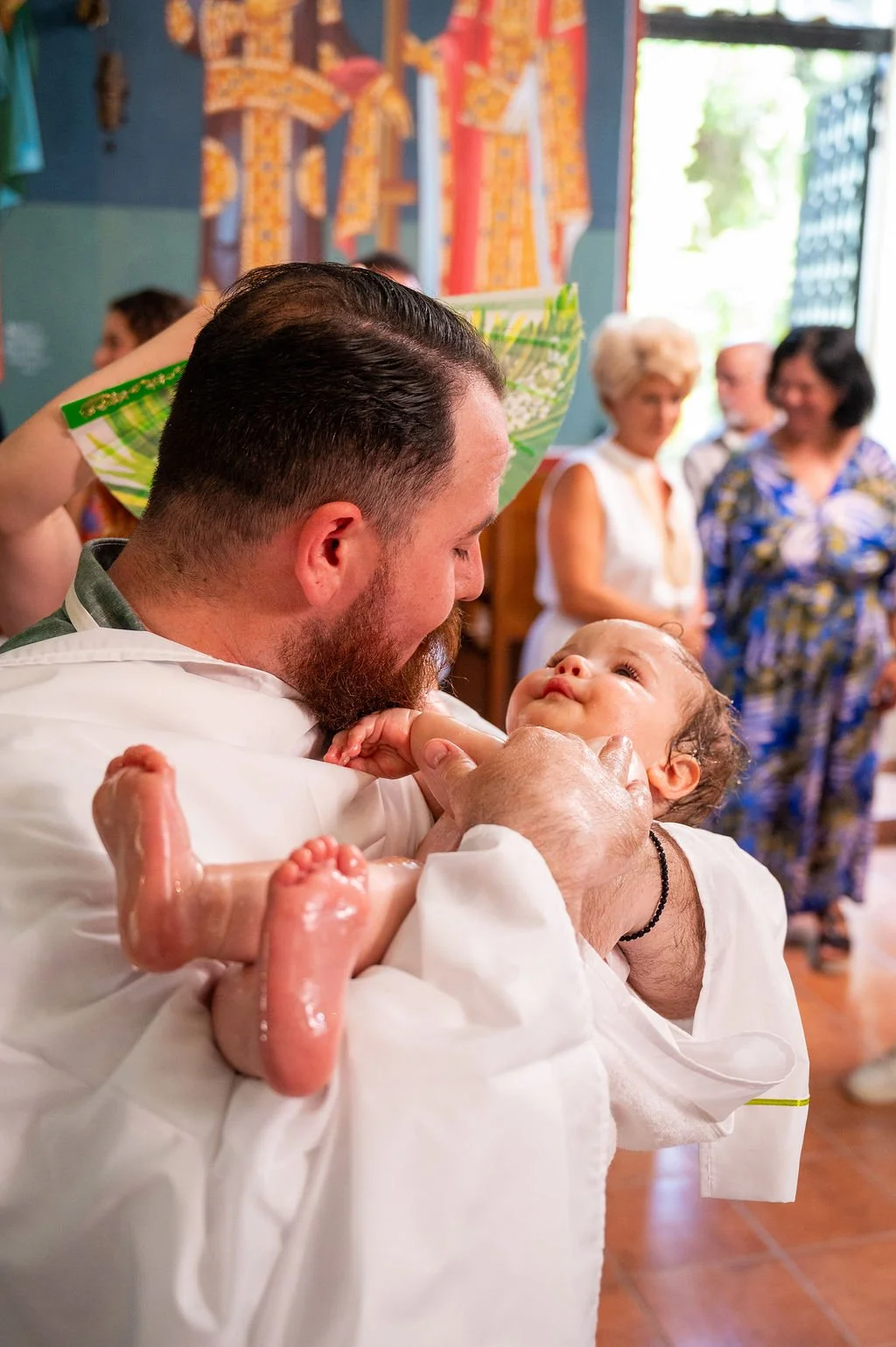A man holding a baby during a baptism ceremony in a colorful room with people in the background.