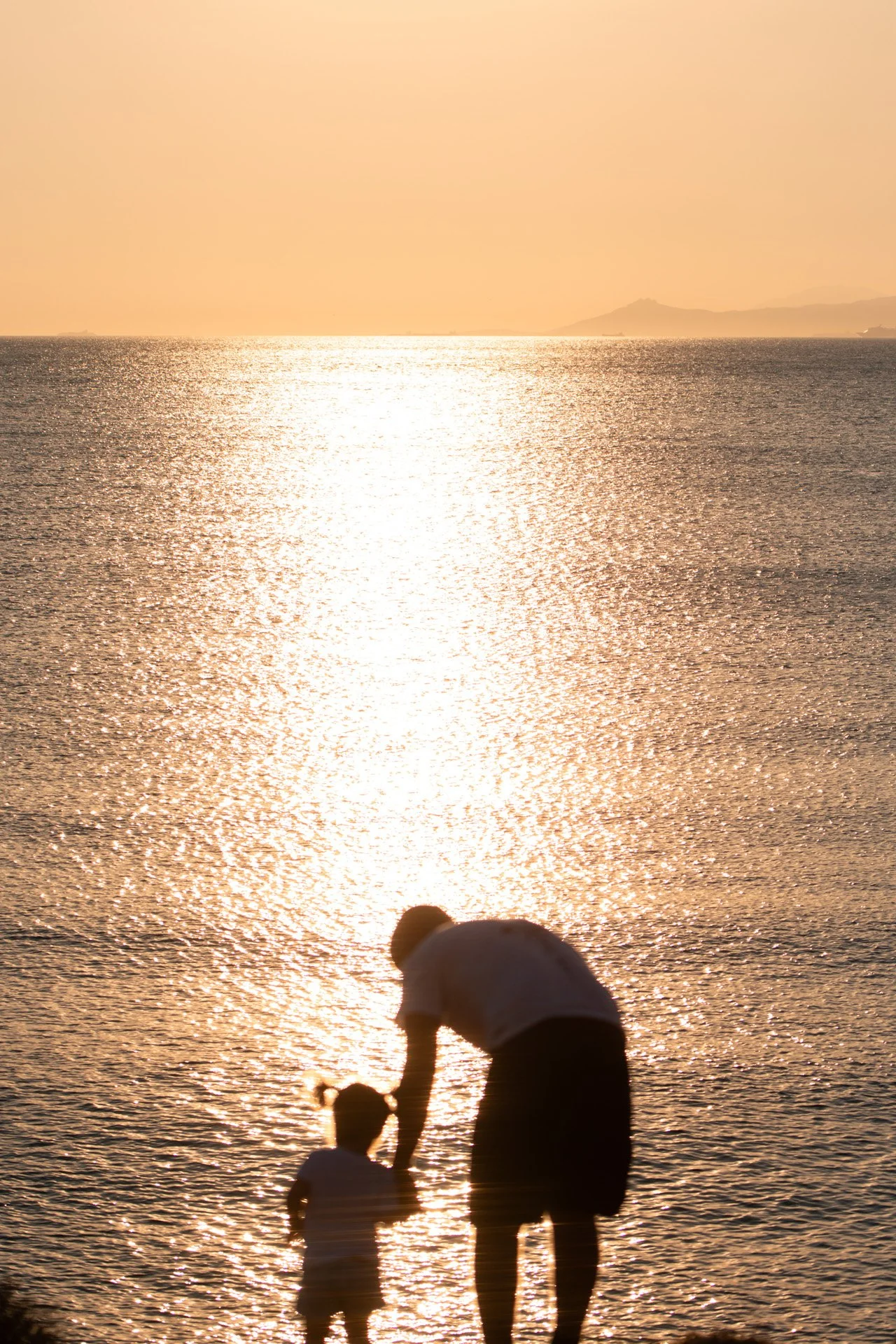 A silhouette of an adult and a child standing by the water at sunset, with a shimmering reflection on the sea surface and distant mountains on the horizon.