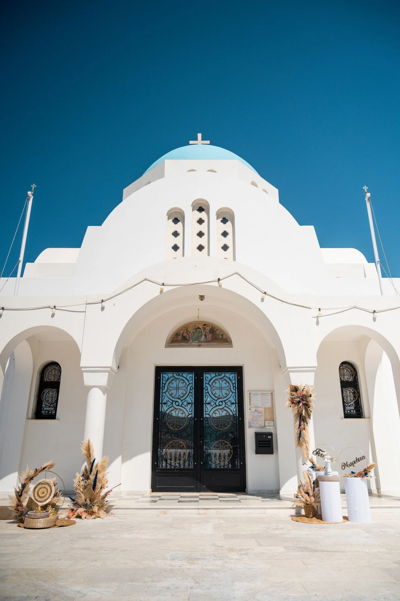 White church with blue dome and cross on top, decorated for a celebration with dried flowers and signs at the entrance, against a clear blue sky.