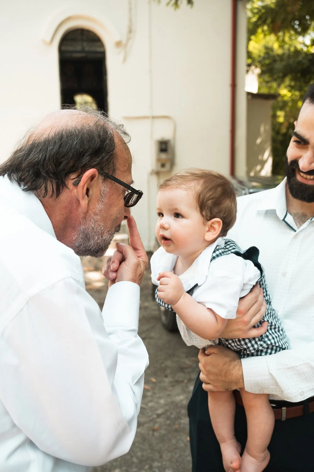 A doctor making a shushing gesture while talking to a young boy, held by a man, outdoors with a building and trees in the background.